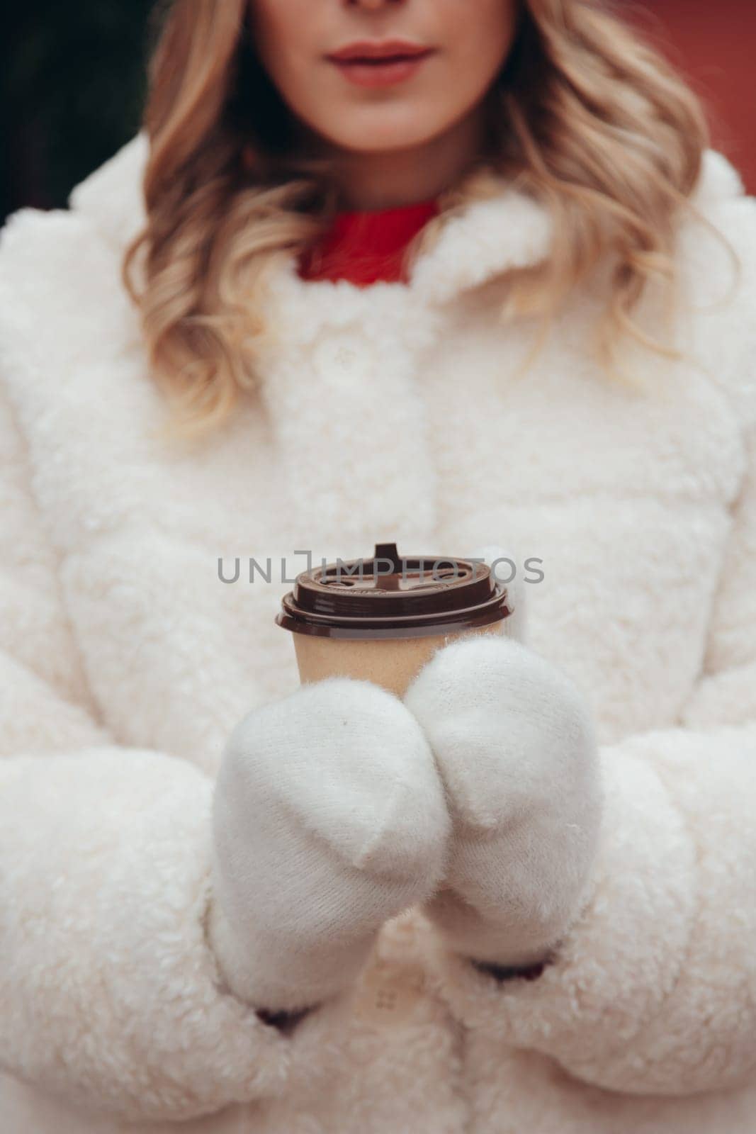 Woman in white wool mittens and fur coat holding take away coffee cup outdoors, soft focus on hands.