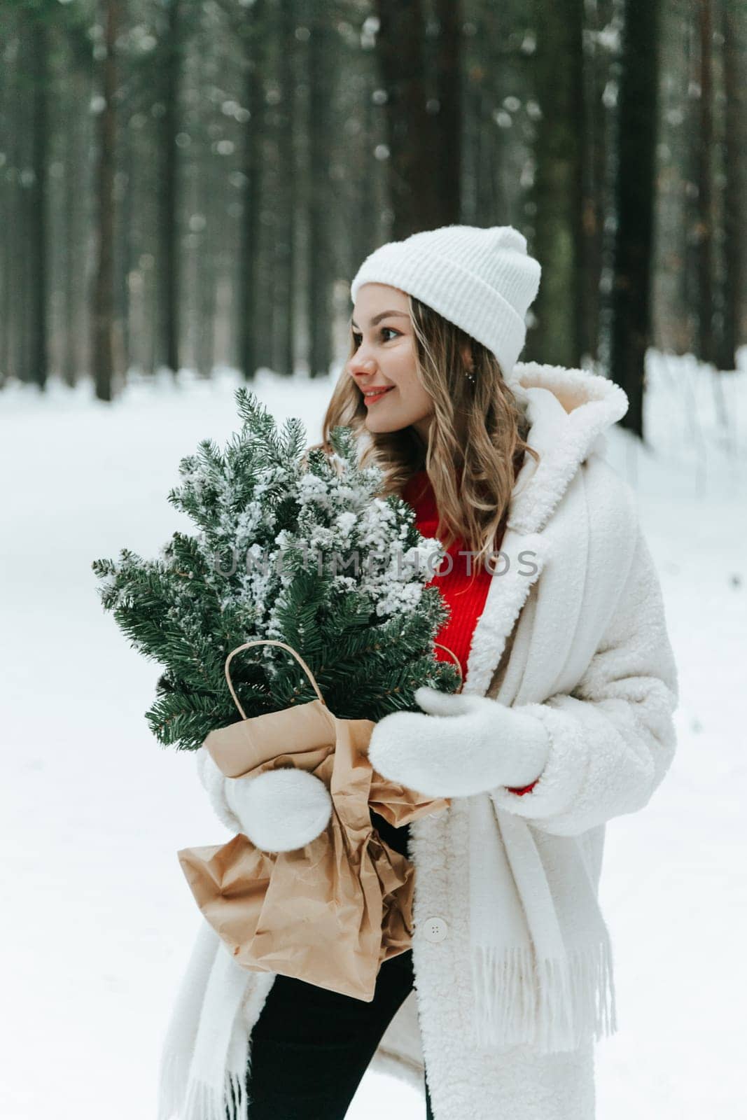 Young blonde woman wears white fur coat, knitted hat and mittens, posing with fir tree branches wrapped with craft paper in winter forest. Winter fashion