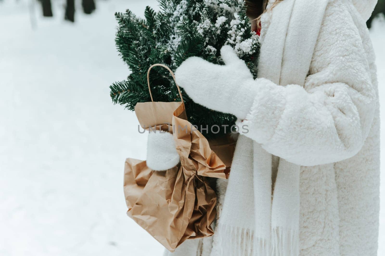 Young blonde woman wears white fur coat, knitted hat and mittens, posing with fir tree branches wrapped with craft paper in winter forest. Winter fashion