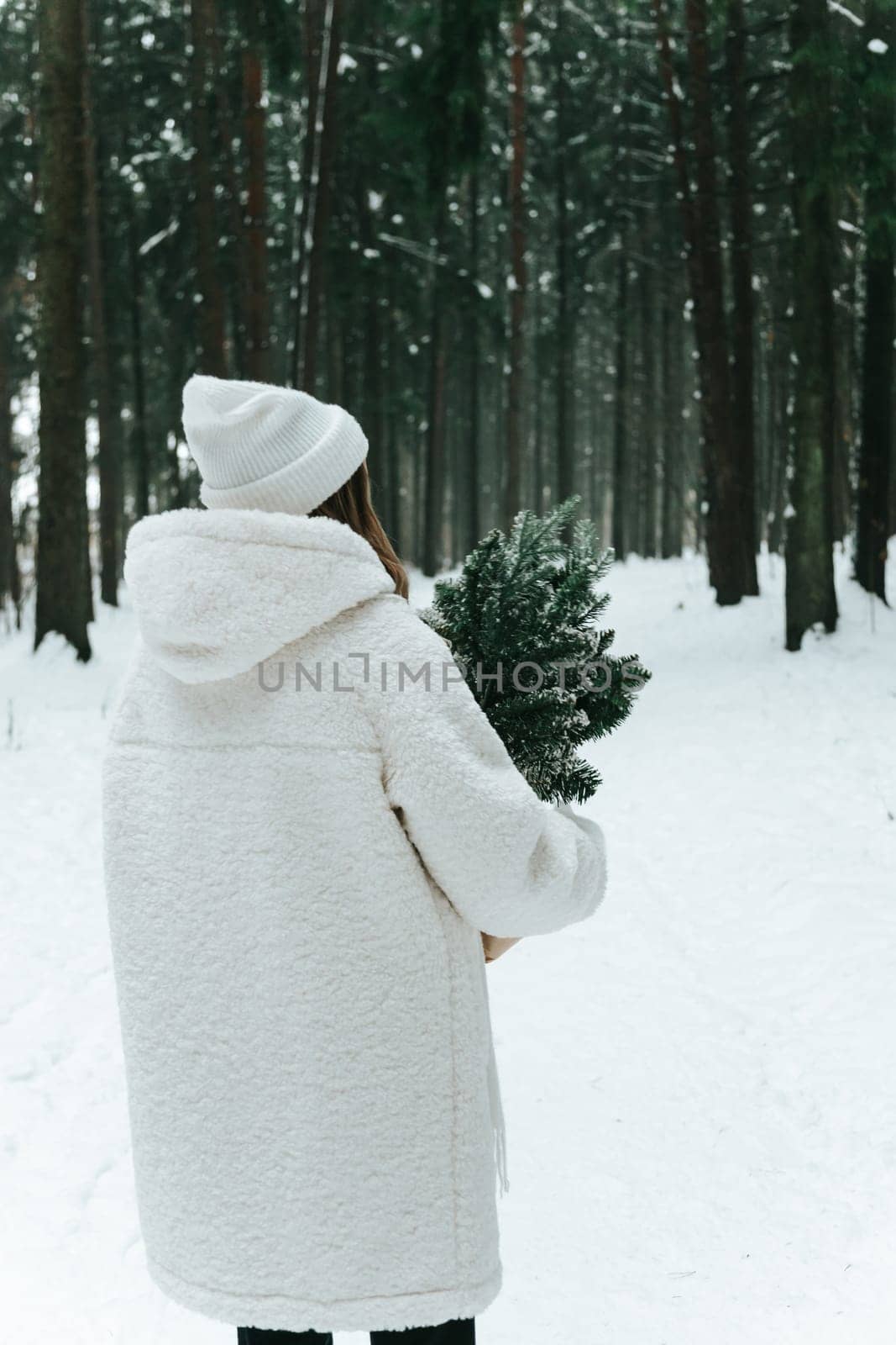Blonde woman wears white fur coat and knitted hat posing with fir tree in winter forest, back view