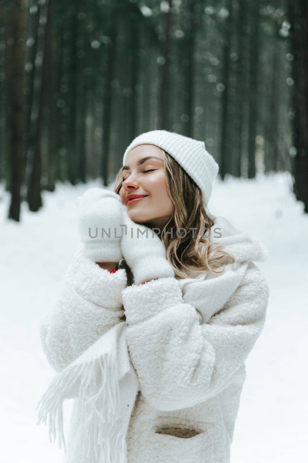 Young woman Enjoying snowy winter forest and warm clothes. Dressed white coat scarf hat and mittens fold palms to face.