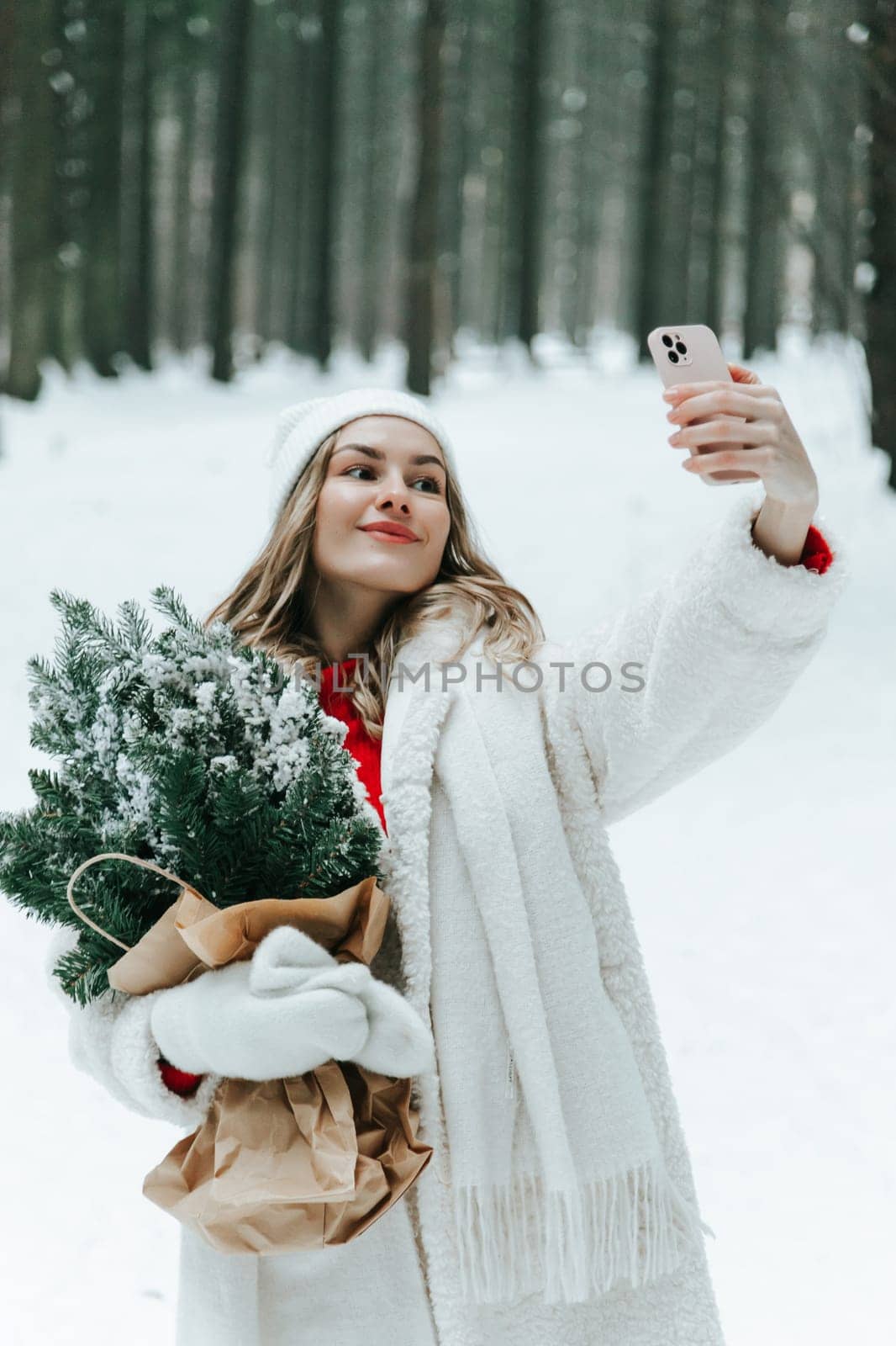 Beautiful girl in a white sweater and a hat with a Christmas tree in her hands makes a selfie in the winter forest.