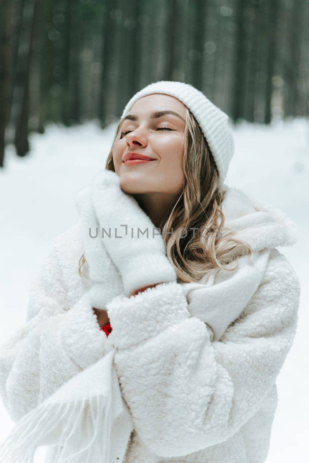Young woman Enjoying snowy winter forest and warm clothes. Dressed white coat scarf hat and mittens fold palms to face.