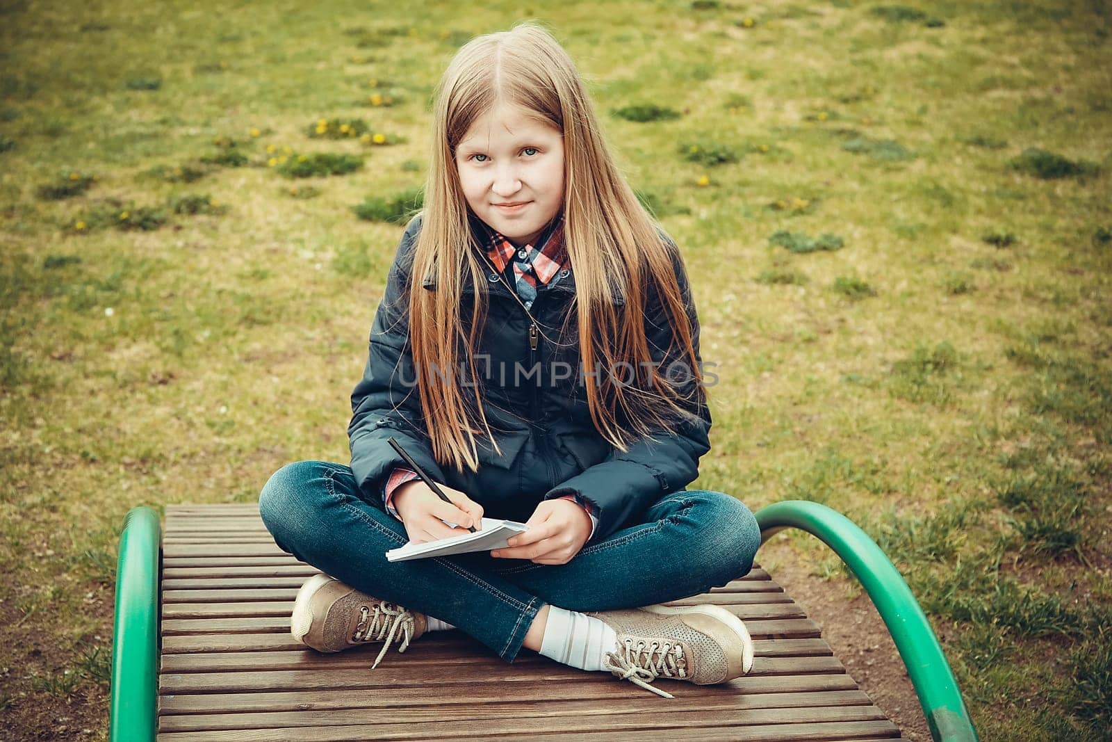 A blond school-age girl sits on a wooden bench in the park during the day and writes notes in a notebook
