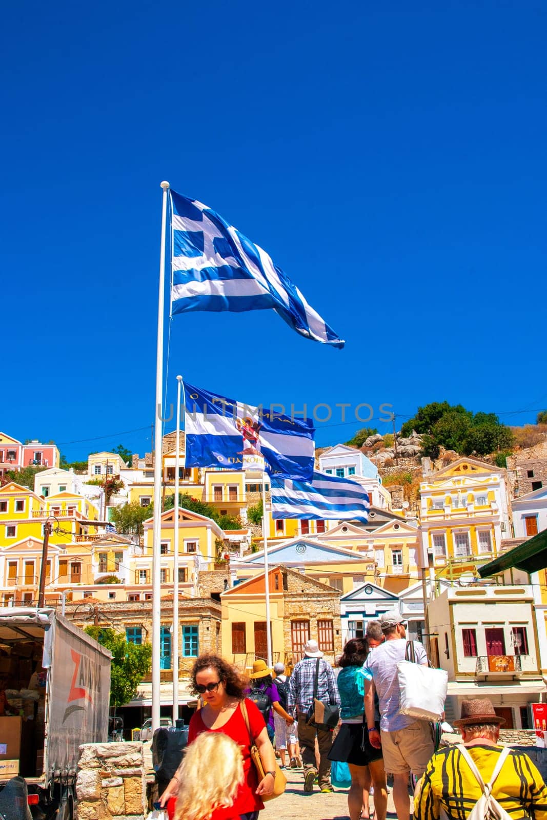 SYMI, Greece - June 03, 2021. Tuorists at the harbour of Symi Town. Colorful villas stretch up the steep slopes.