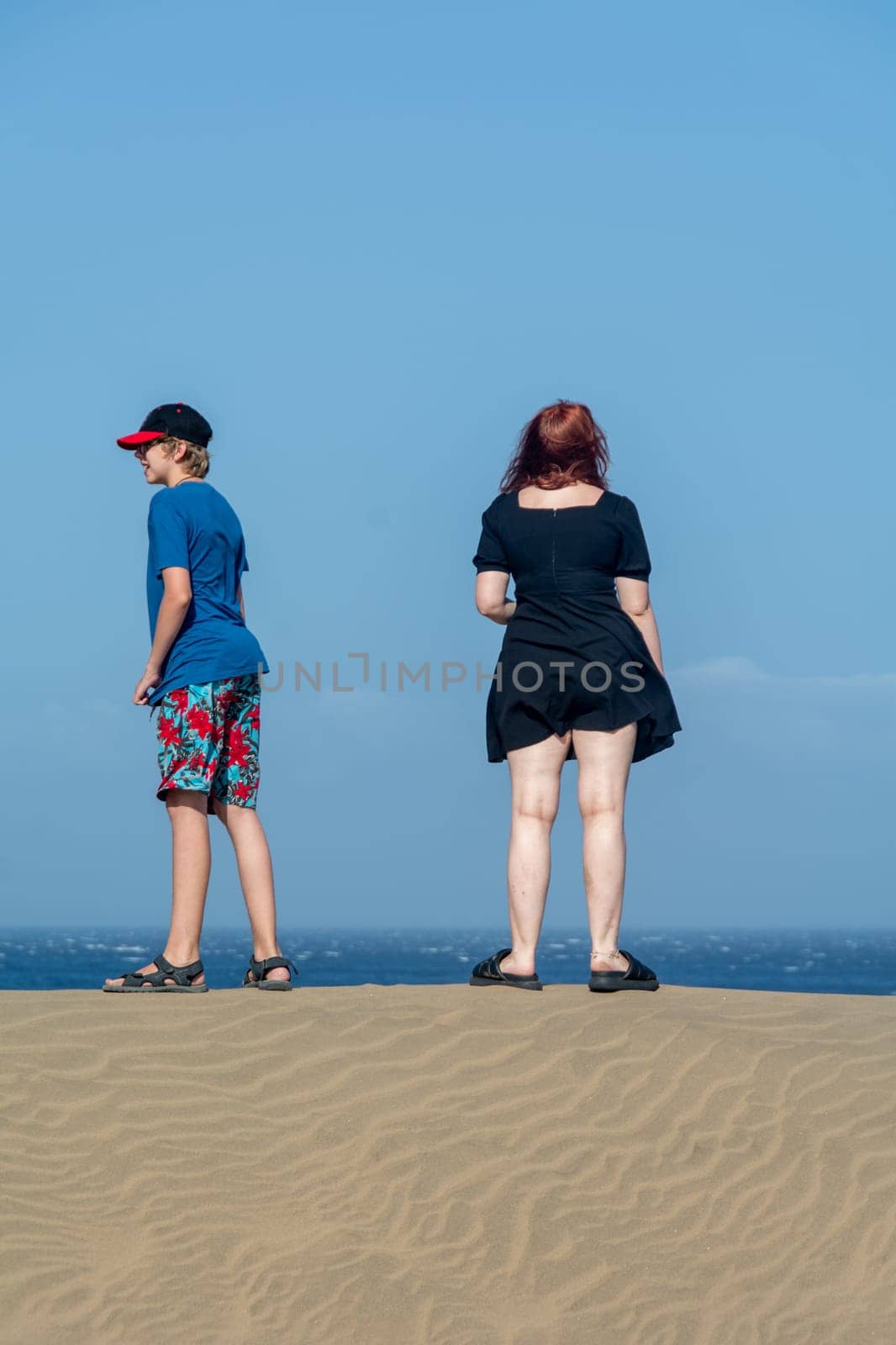 Two teenagers stand on a sand dune, with sea and blue sky
