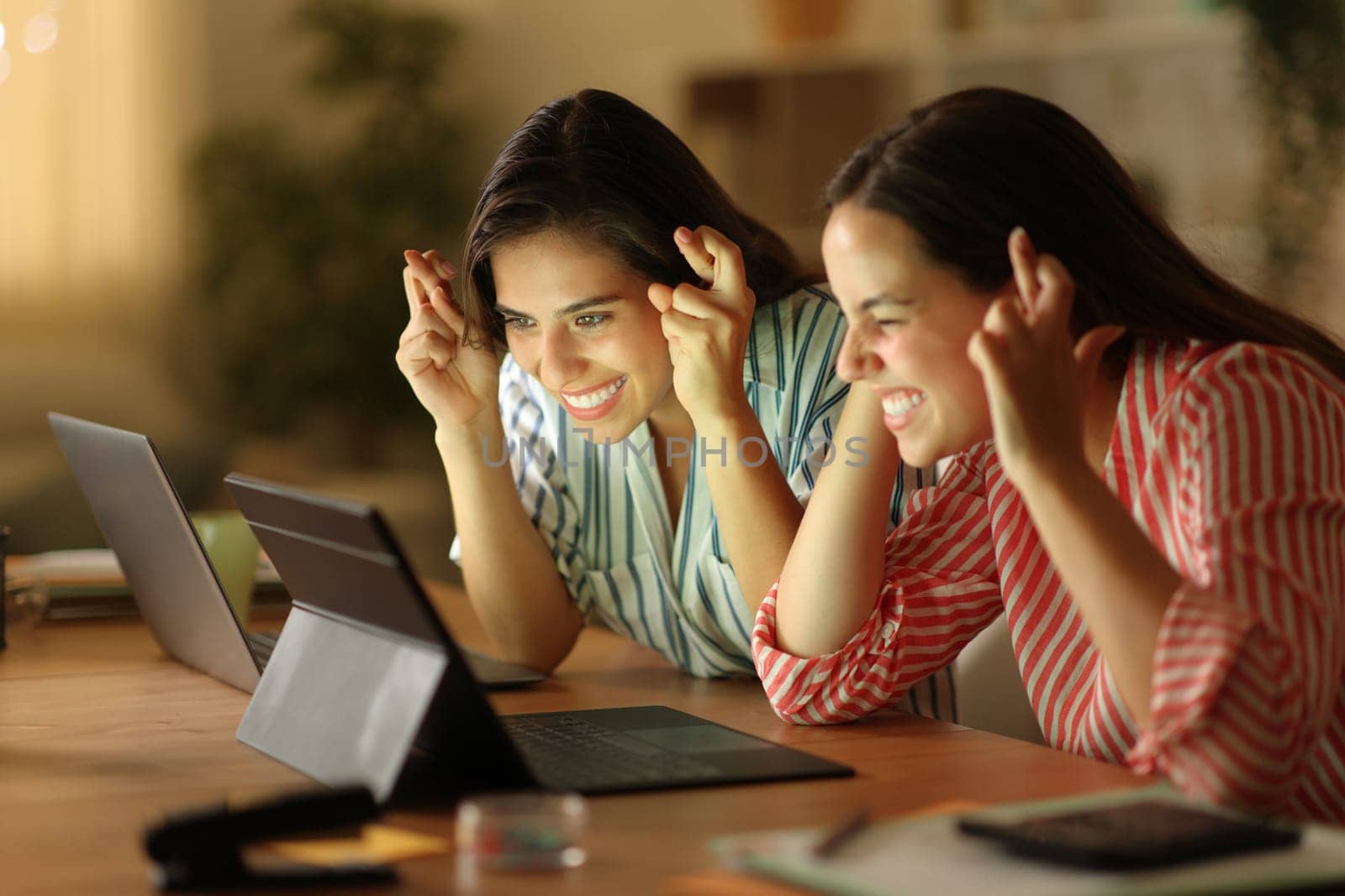 Two tele workers crossing fingers waiting for online result checking laptop in the night
