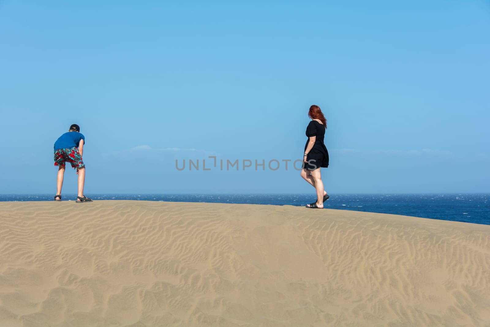 Two teenagers stand on a sand dune, with sea and blue sky