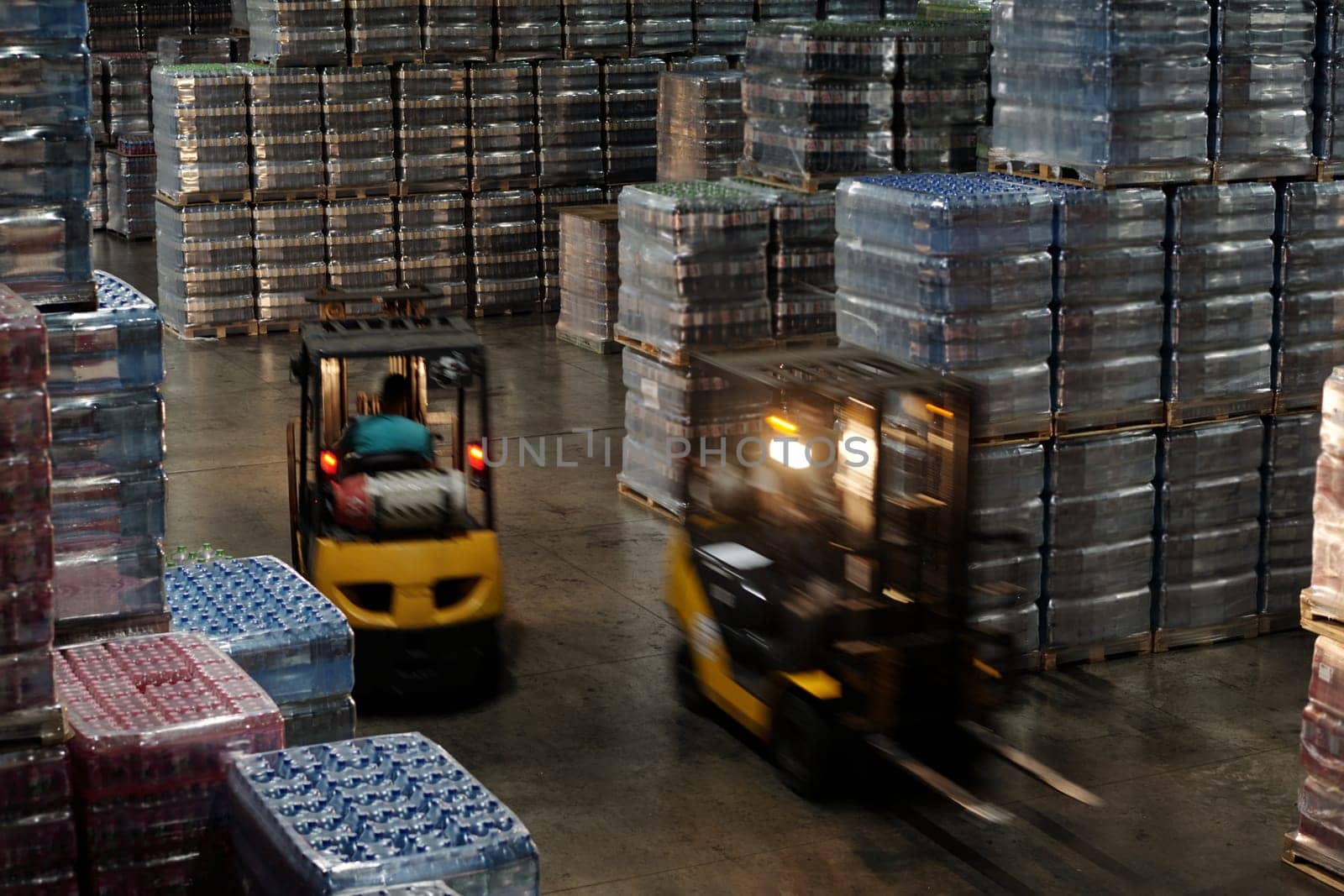 Blurry motion of two forklifts along aisle between huge stacks of packed plastic bottles containing mineral water, soda or some other drinks
