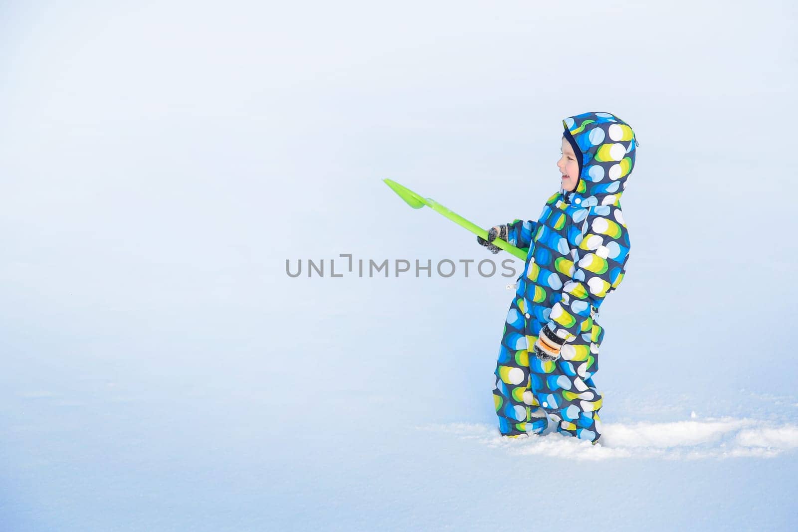 Laughing a little boy in winter with a green shovel in overalls makes his way through a snow-covered field