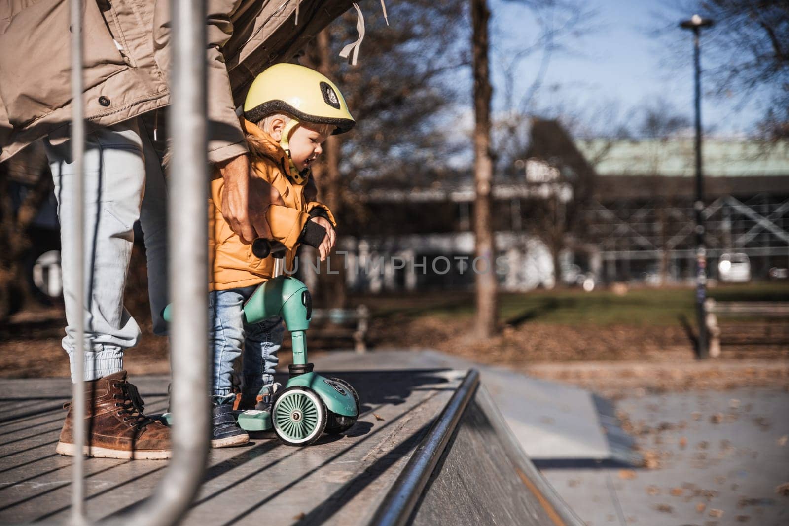 Father supervises his fearless small toddler boy while riding baby scooter outdoors in urban skate park. Child wearing yellow protective helmet.
