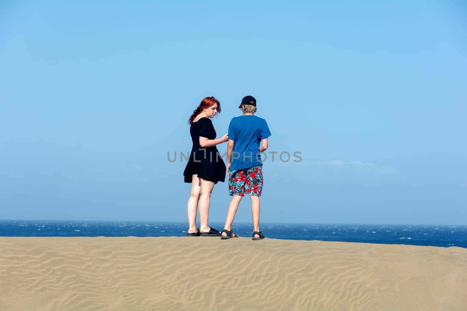 Two teenagers stand on a sand dune, with sea and blue sky