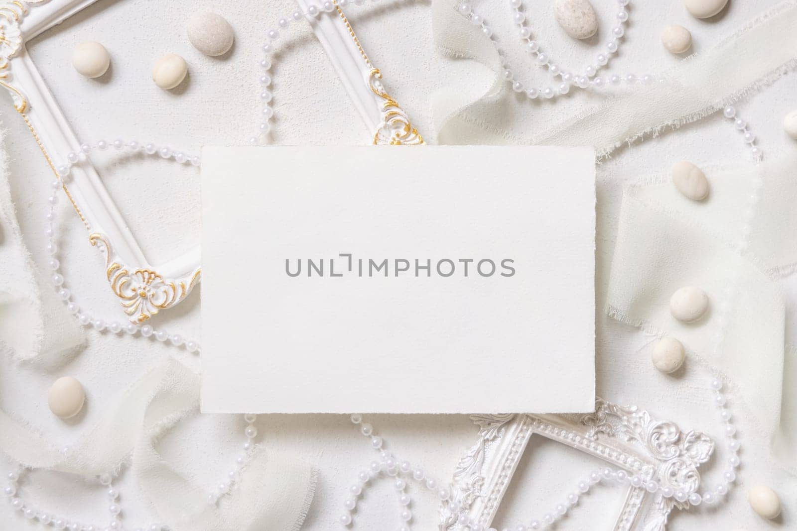 Blank paper card near white frames, pearls, pebbles and silk ribbons on white table top view, wedding mockup. Romantic scene with horizontal card flat lay. Valentines, Spring or Mothers day concept