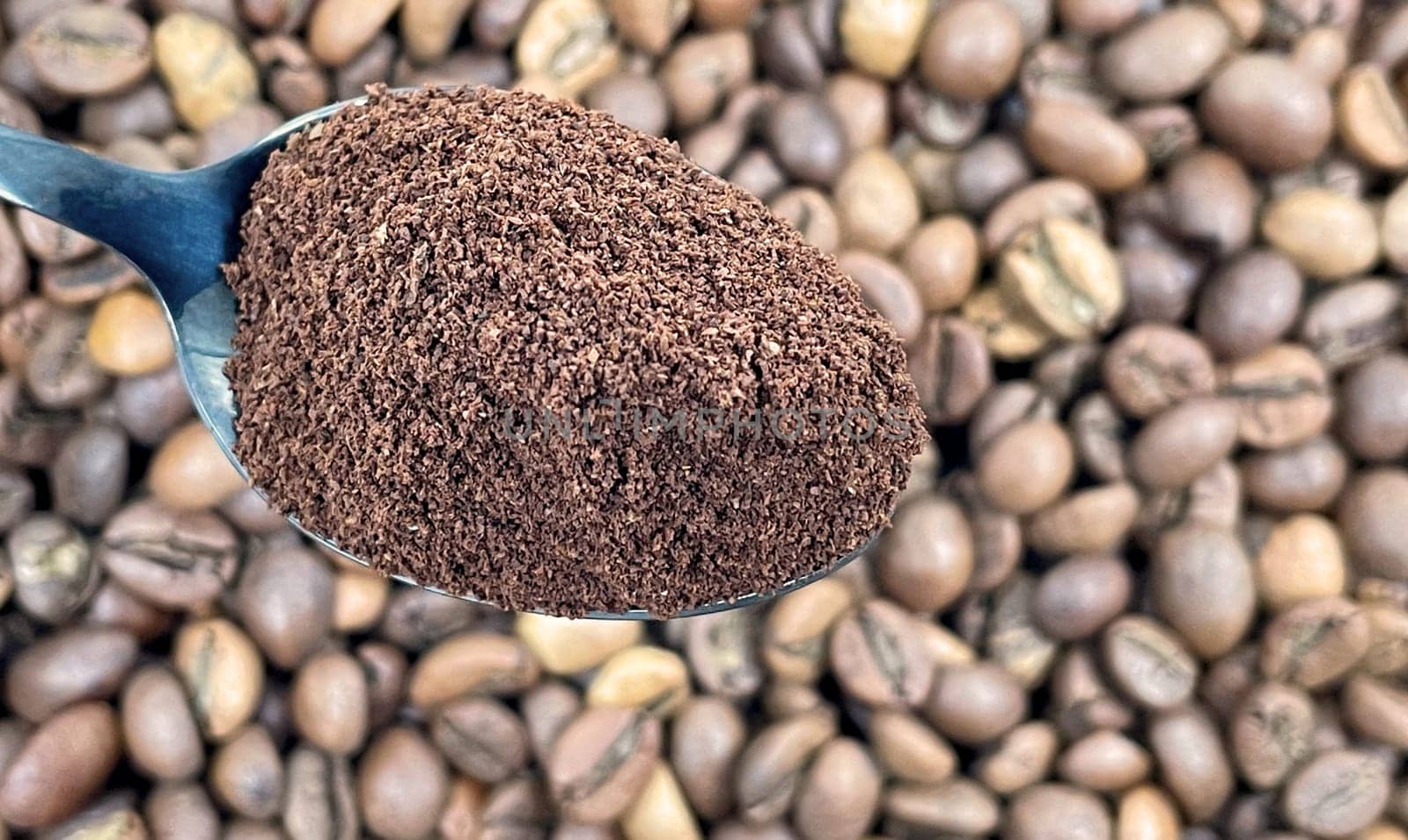 Ground coffee powder in a metal spoon in the foreground, against the background of roasted aromatic coffee beans. Coffee concept