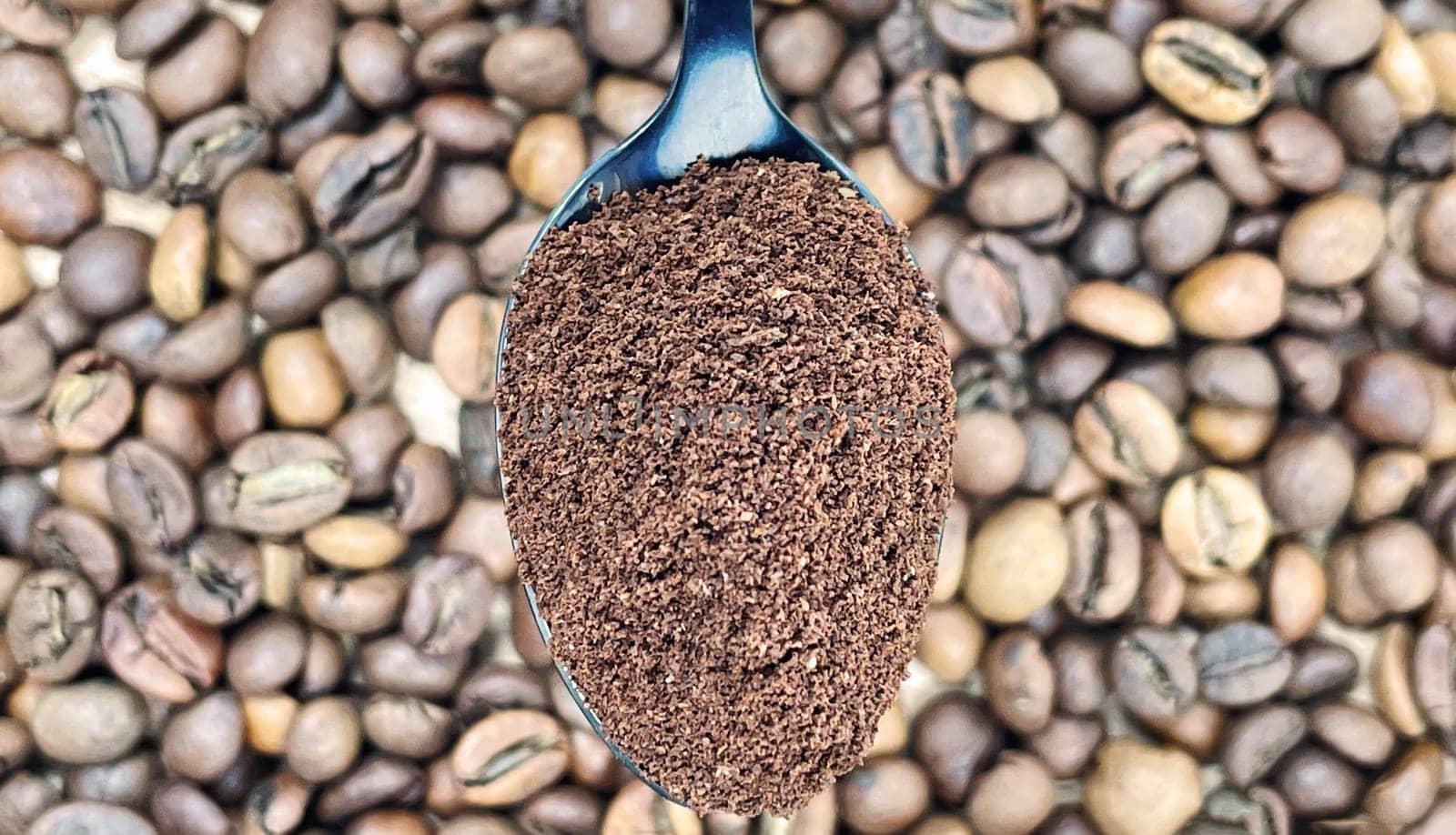 Ground coffee powder in a metal spoon in the foreground, against the background of roasted aromatic coffee beans. Coffee concept