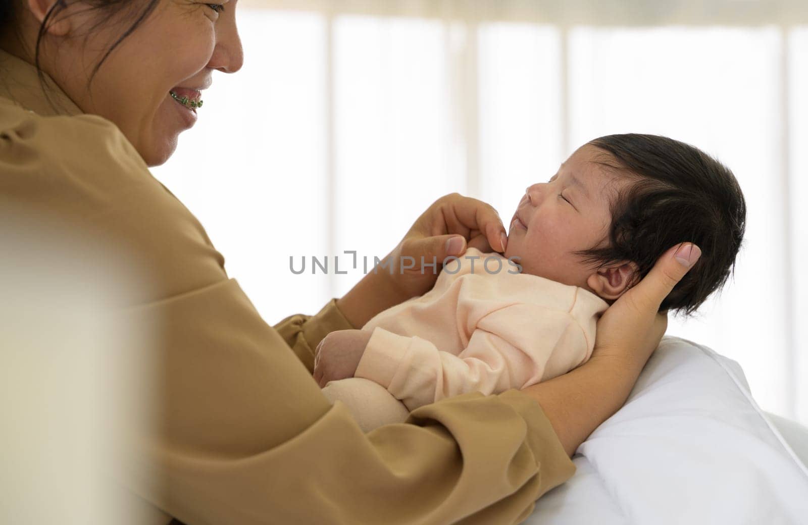 Happy cute newborn baby girl or boy 1 month sleeping on mom hand at home. Smiling mother sitting on white bed and gently touching chin of infant daughter or son with love. Care, love and mothers day concept.