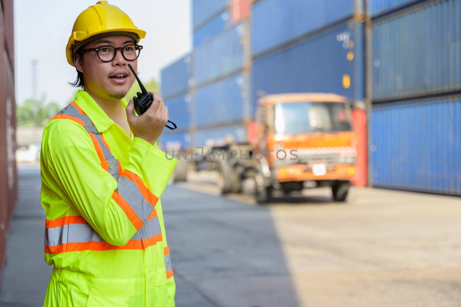 Asian worker in yellow hardhat and safety vest using walkie talkie in cargo shipping yard for communication with driver on loading containers