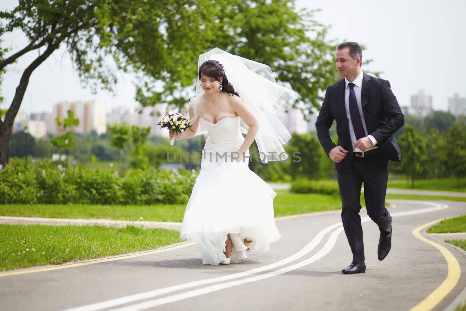 The bride and groom run together along an asphalt road on a summer day