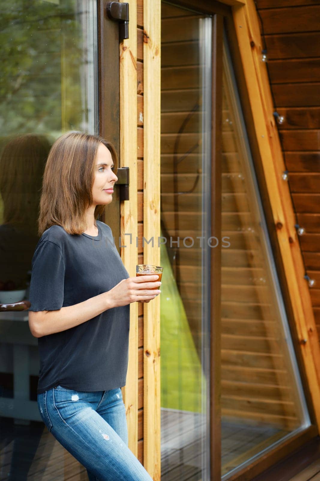 Cute woman came out of a wooden house, leaned against a glass door and drinks juice from a glass
