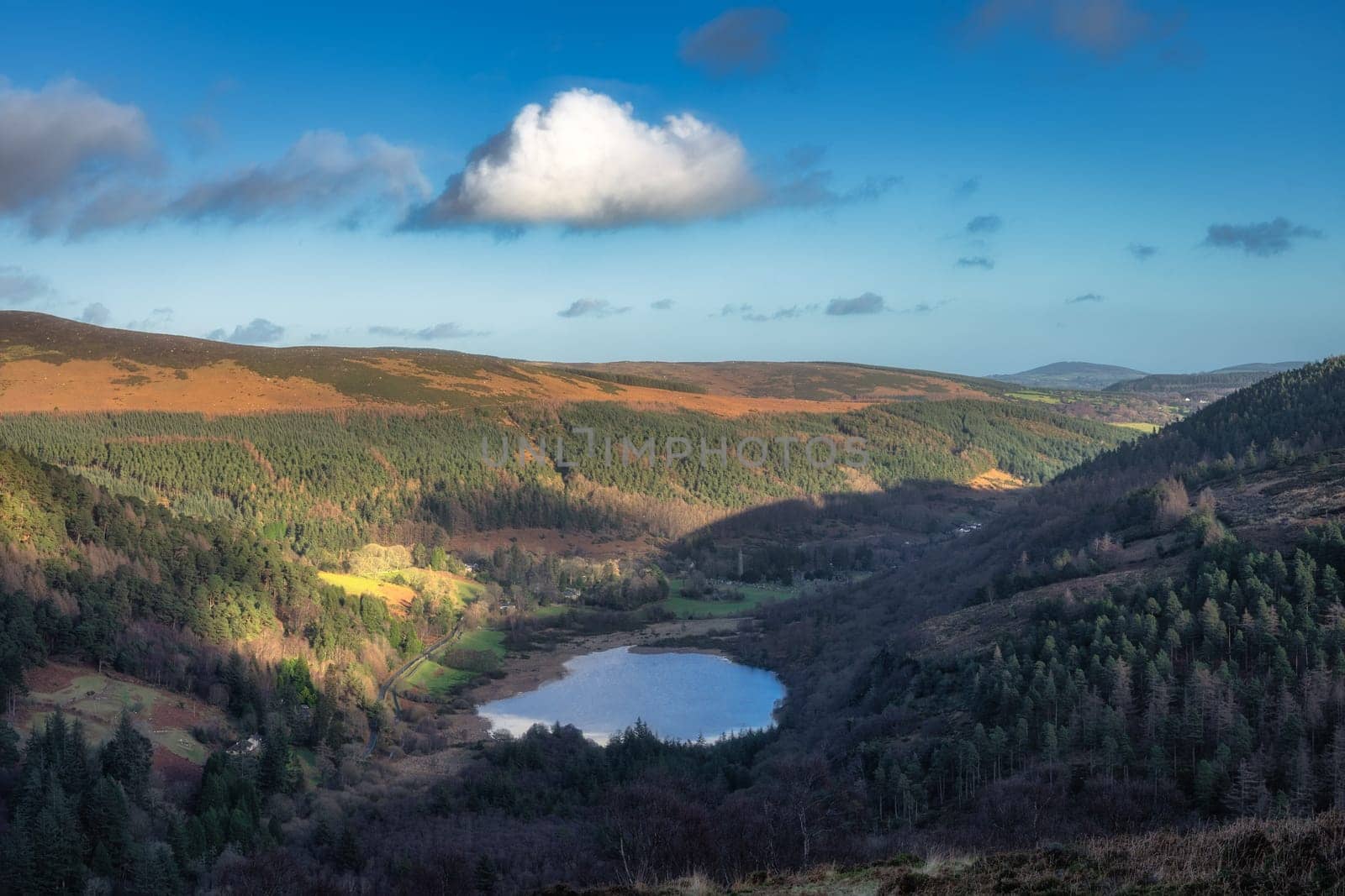 Glendalough lake and cemetery in the valley. Forest on the hills illuminated by sunlight. Hiking in beautiful autumn Wicklow Mountains, Ireland