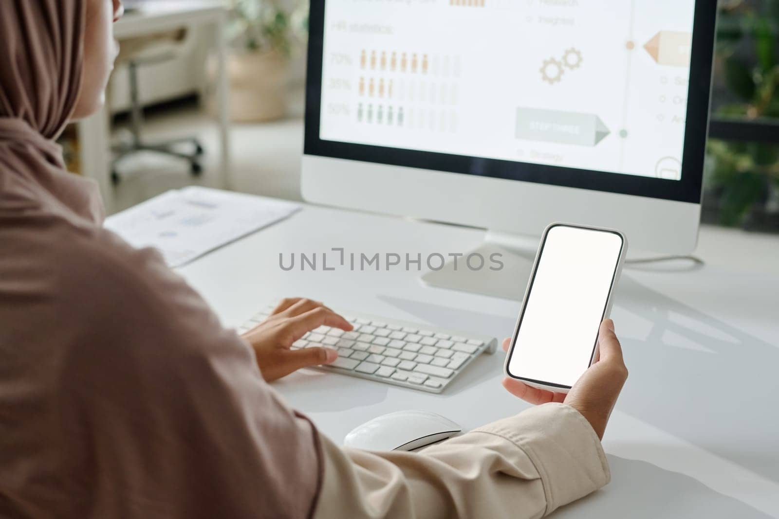 Smartphone with blank screen held by young Muslim businesswoman in hijab sitting by workplace in front of computer and typing
