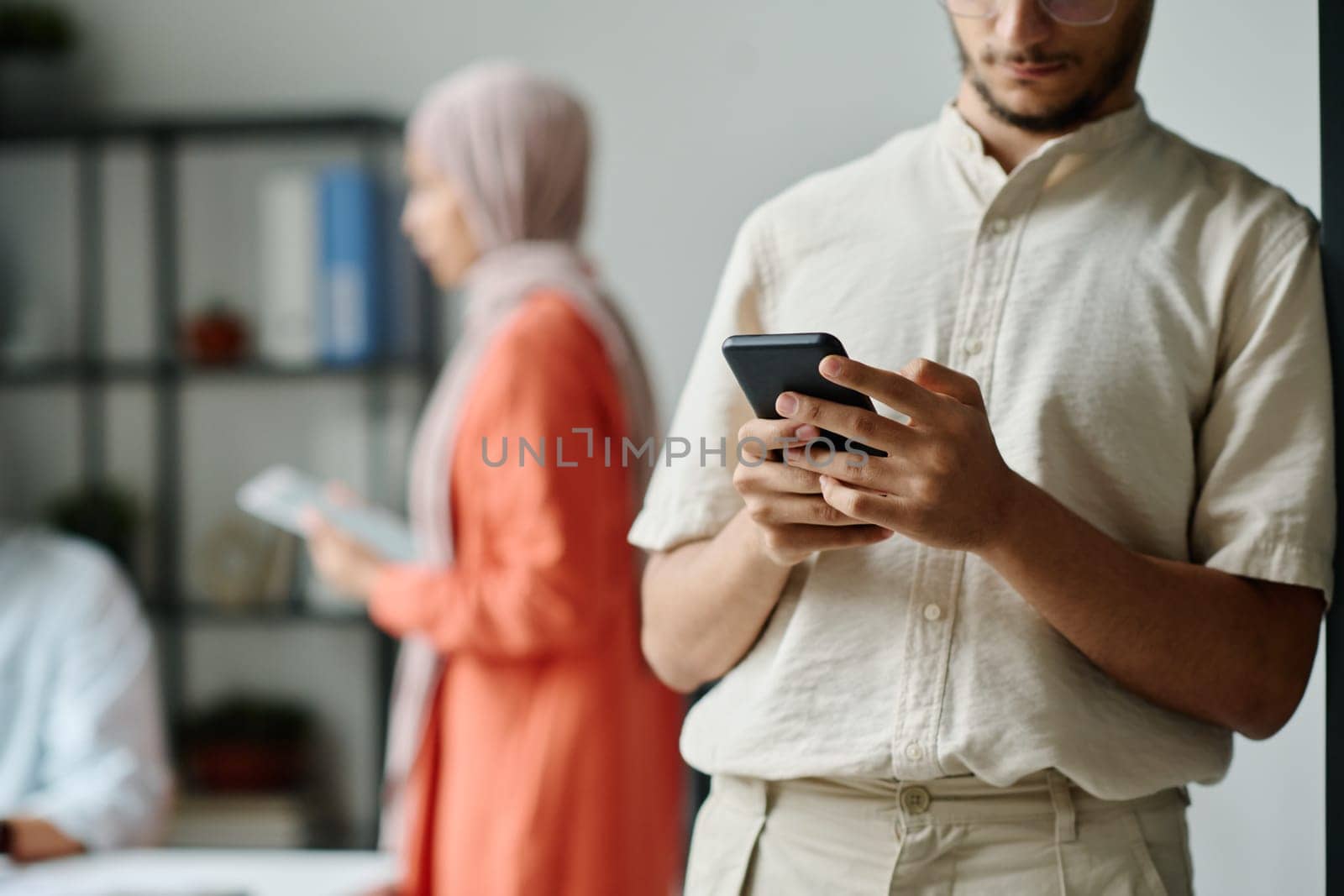Cropped shot of young Muslim businessman in casualwear standing in front of camera and texting in mobile phone in coworking space