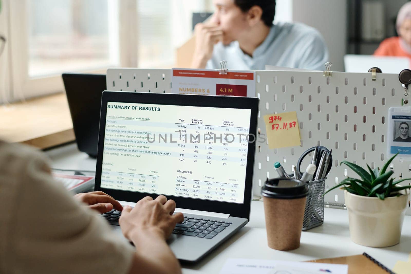Financial data on screen of laptop being used by young male economist sitting in front of pensive colleague looking through office window