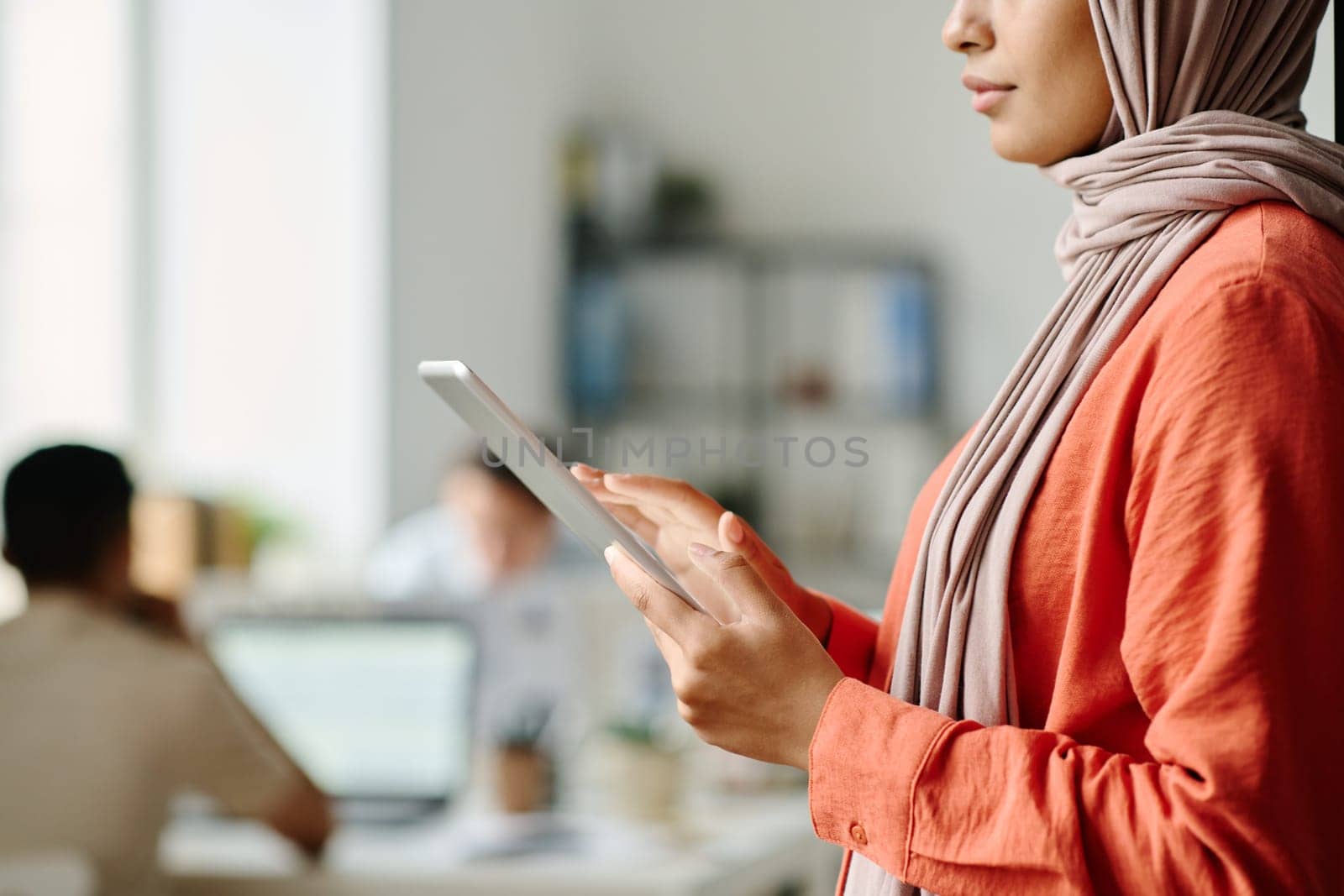 Cropped shot of young Muslim female manager using tablet while standing in coworking space against male colleagues