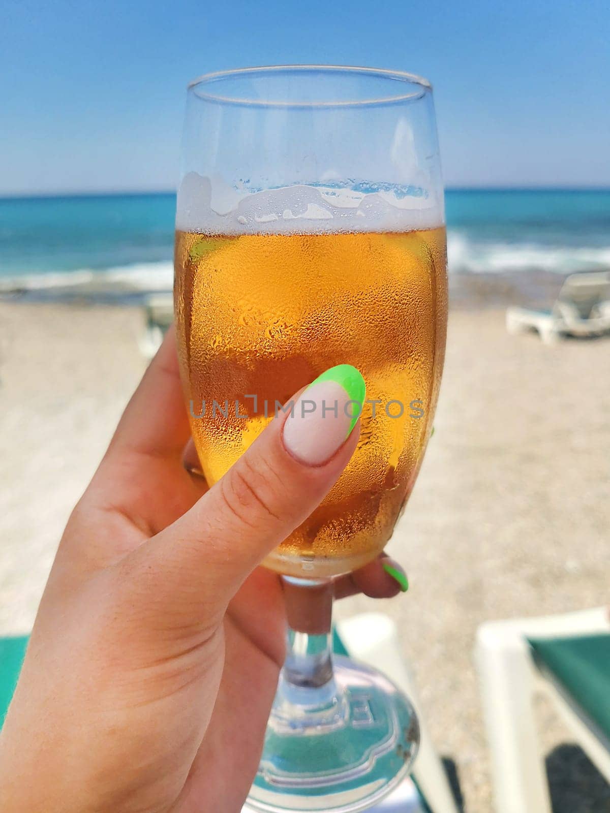 The hand of a girl with a beautiful manicure holds a glass of cold beer on the background of the sea