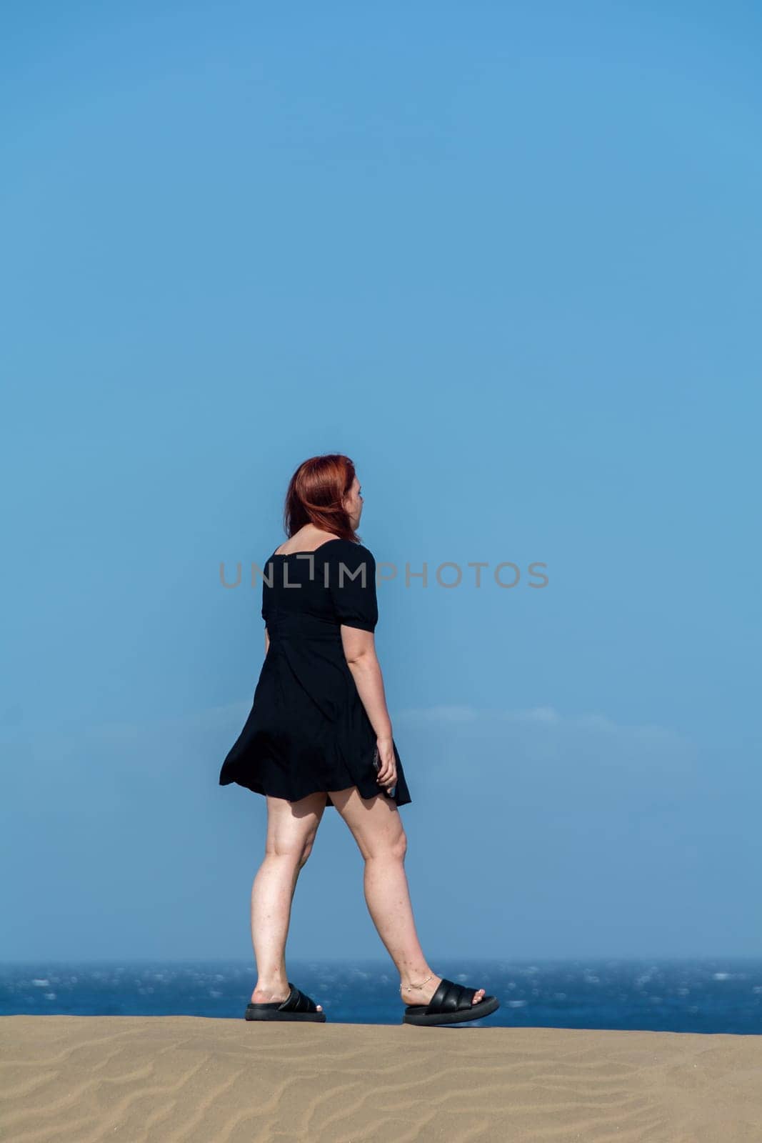 Young girl  in a black  short summer dress stands on a sand dune with a blue sky