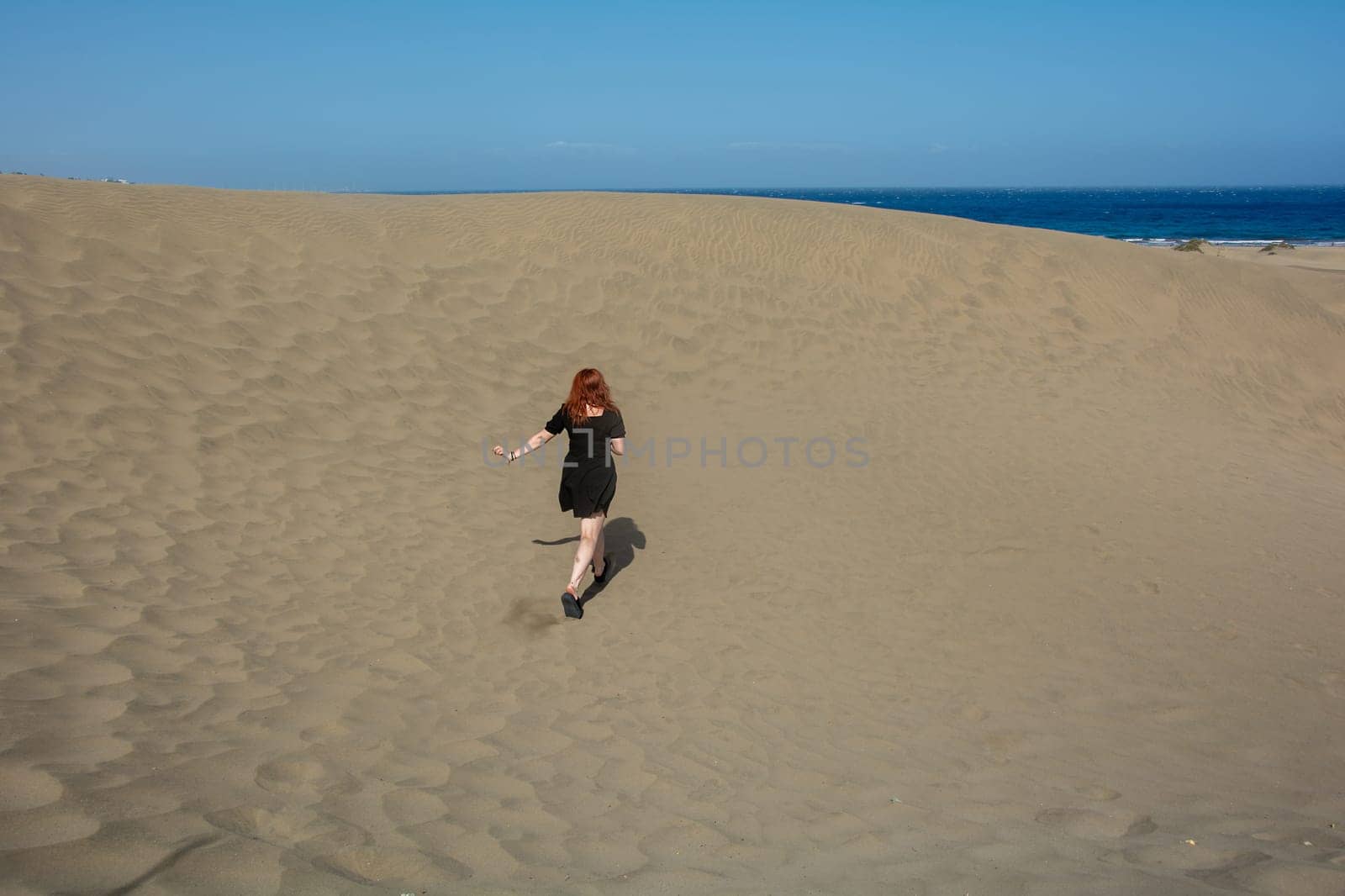 Young woman running up a sand dune towards the sea, with blue sky