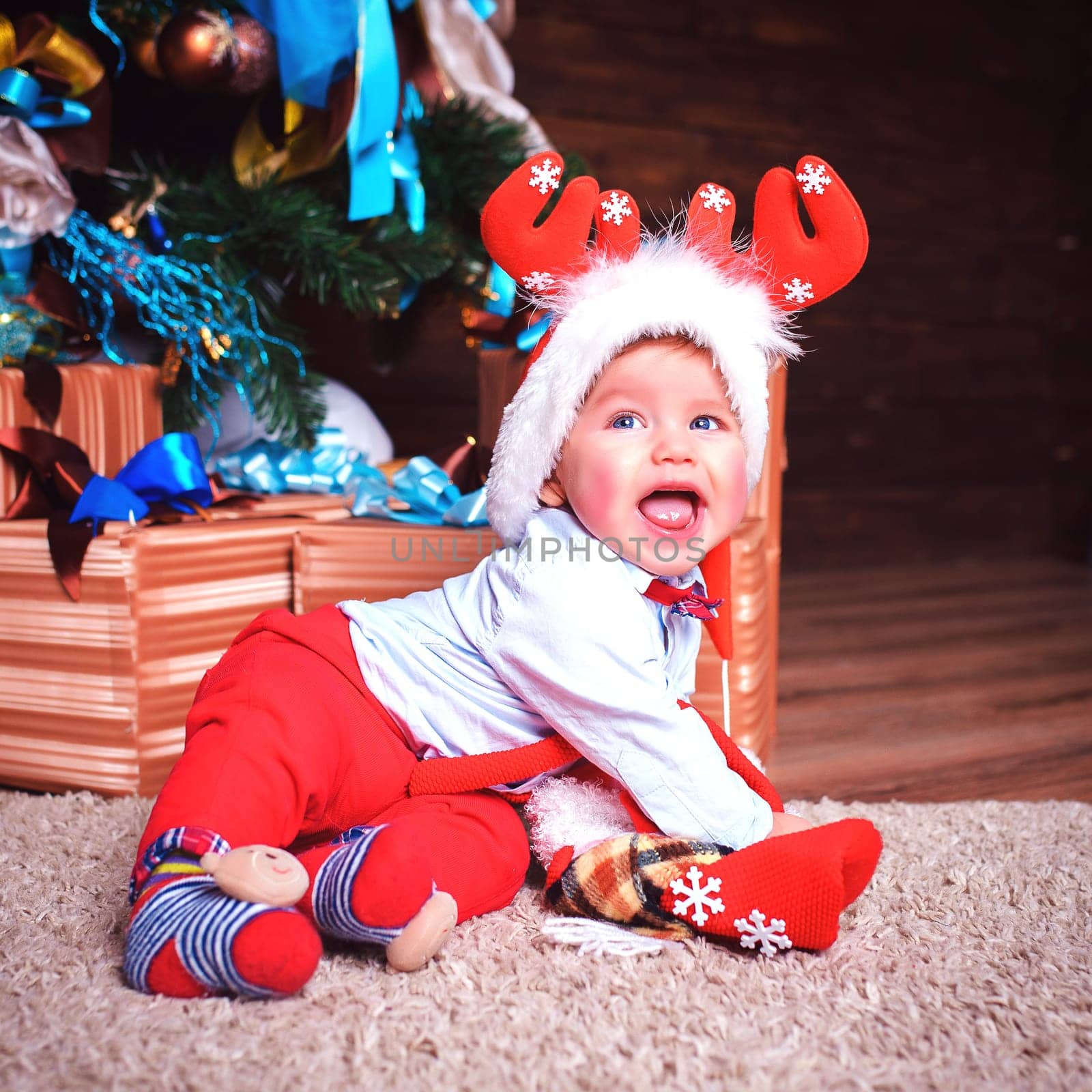 Laughing a little boy with deer antlers on his head is sitting in the studio near the Christmas tree and a bunch of gifts