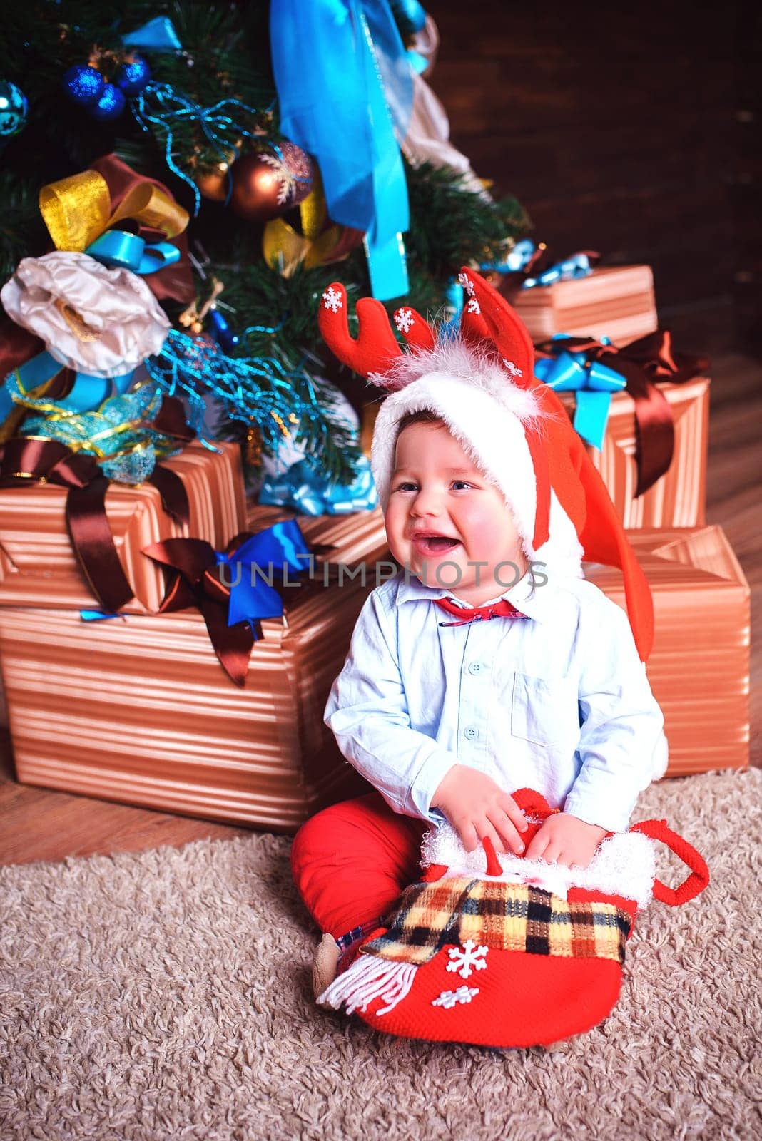 Laughing a little boy with deer antlers on his head is sitting in the studio near the Christmas tree and a bunch of gifts