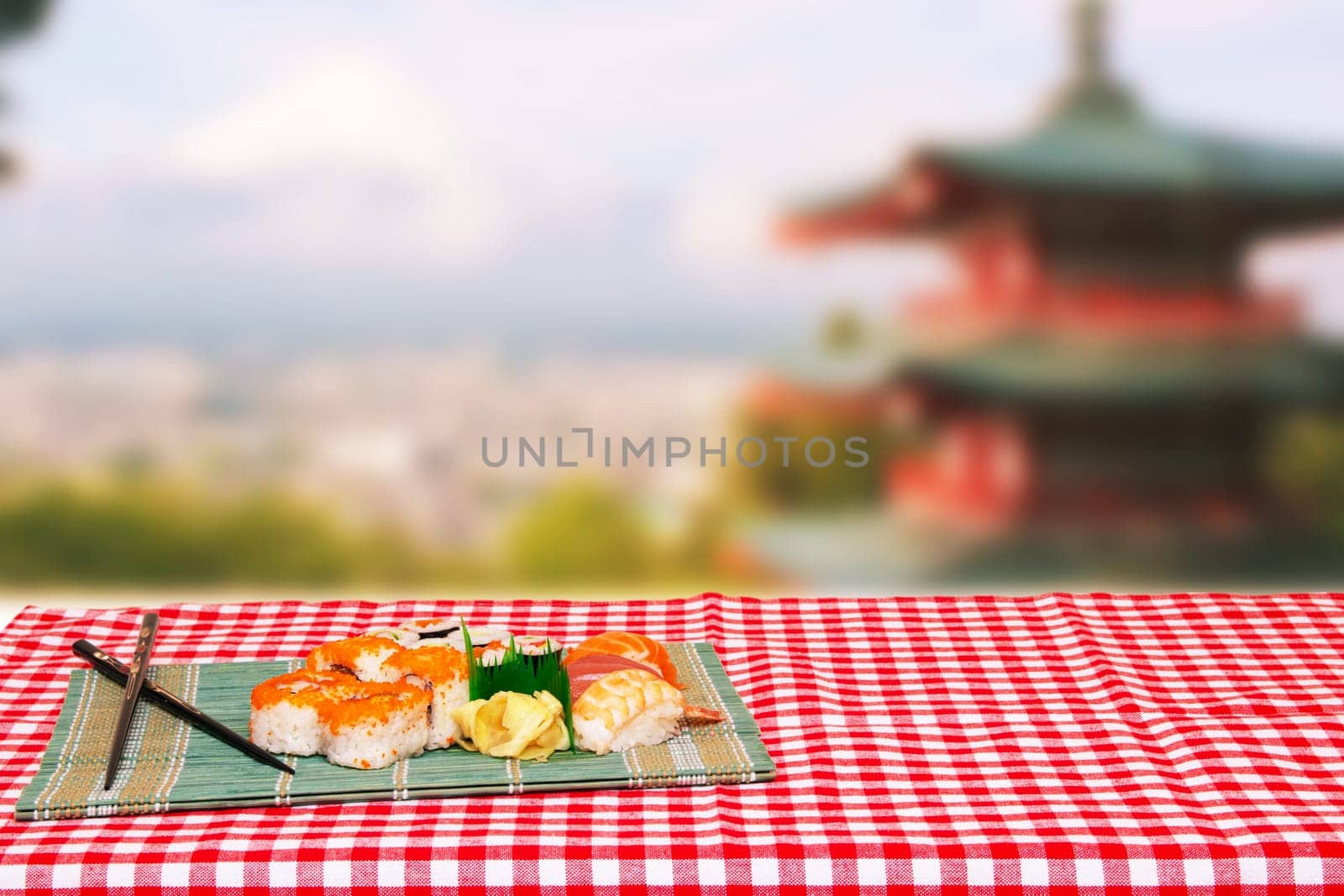 Table top on sushi background. Close-up of various kinds of sushi and chopsticks served on green bamboo mat on a red checkered tablecloth over abstract blurred japanese landscape.