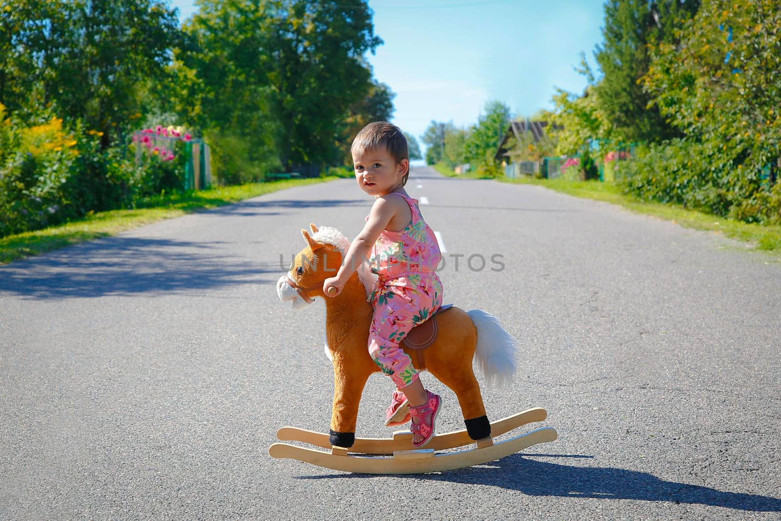 A little girl swings on a toy wooden horse outdoors in the middle of the road on a summer day