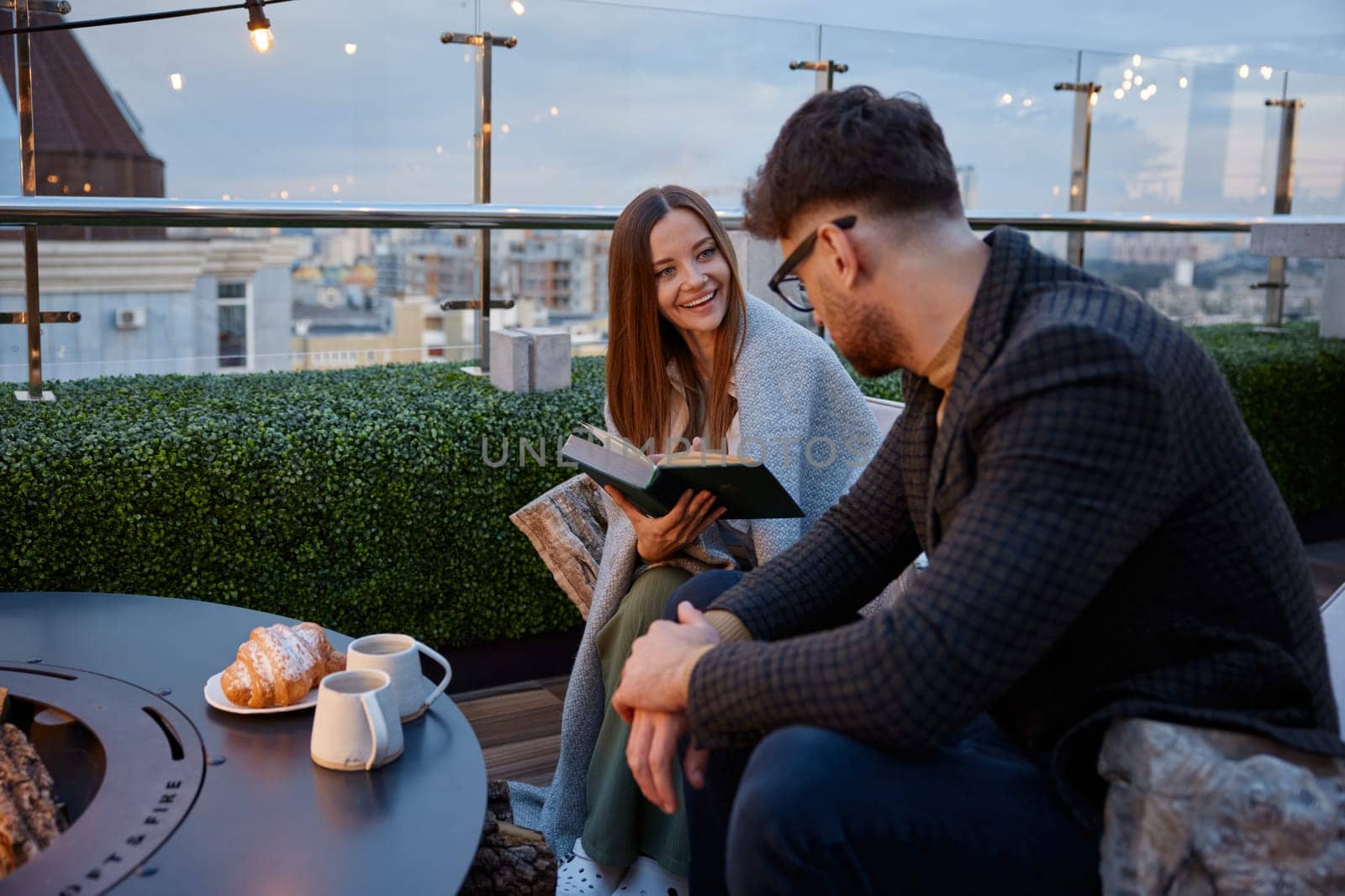 Young girlfriend reading for boyfriend while couple resting at roof top cafe. Excited female sharing impression of favorite book. Young family leisure activity in evening