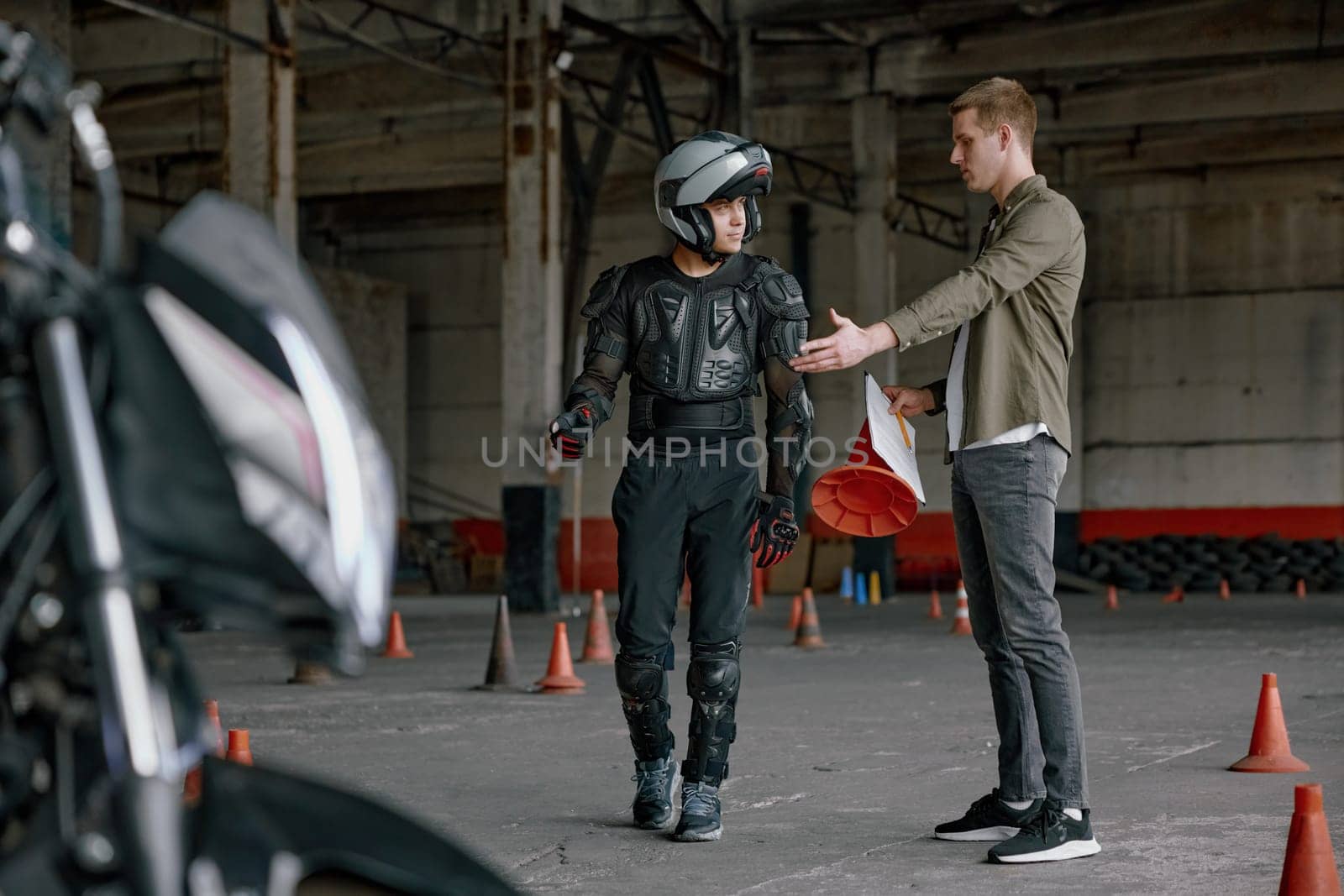 Student motorcyclist and instructor discussing rout of future exam race. Two men standing together looking at checklist at indoor motordrome of driver training center
