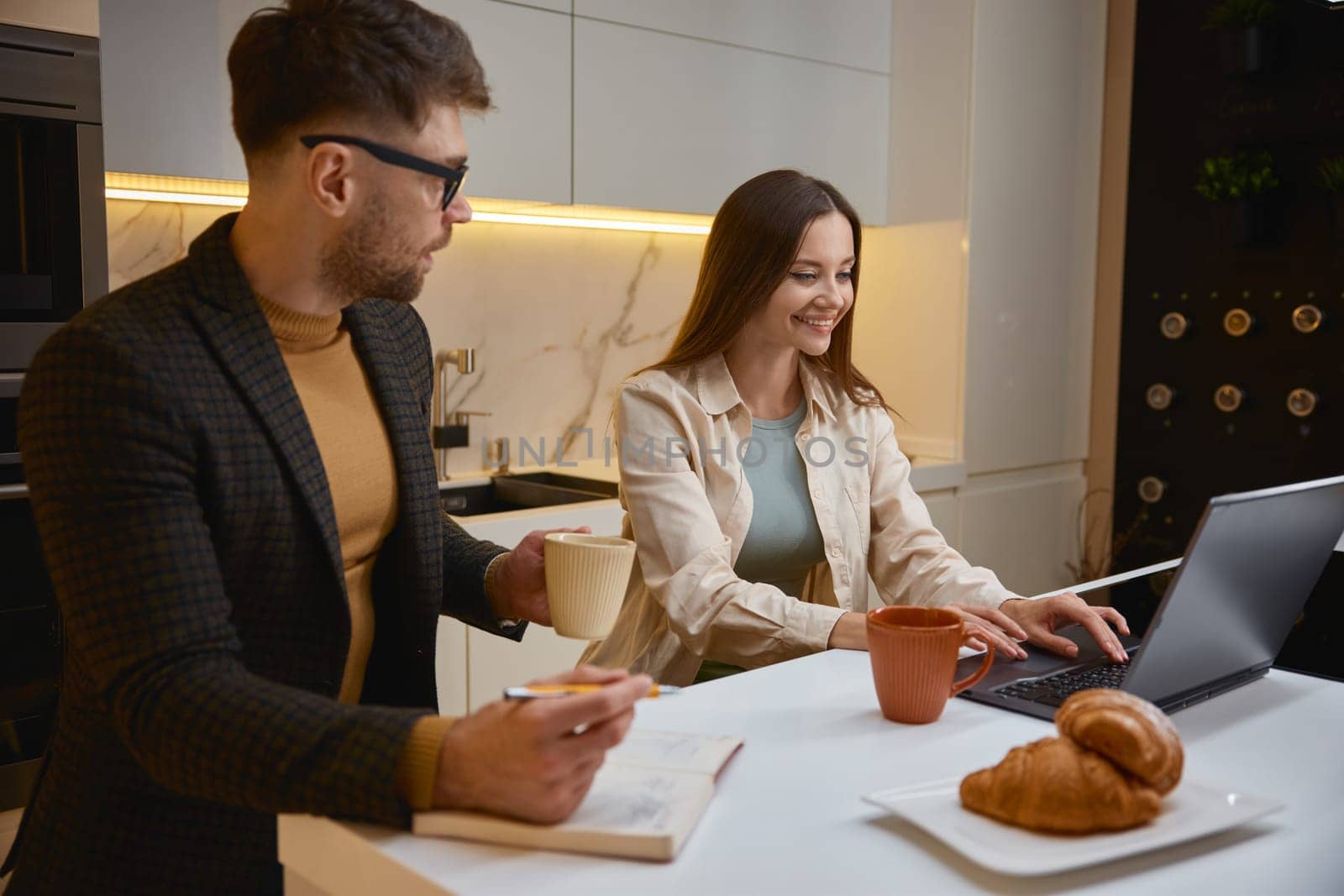 Man and woman freelancer coworkers working with paper document and laptop computer while drinking coffee having snack at office kitchen. Modern coworking space