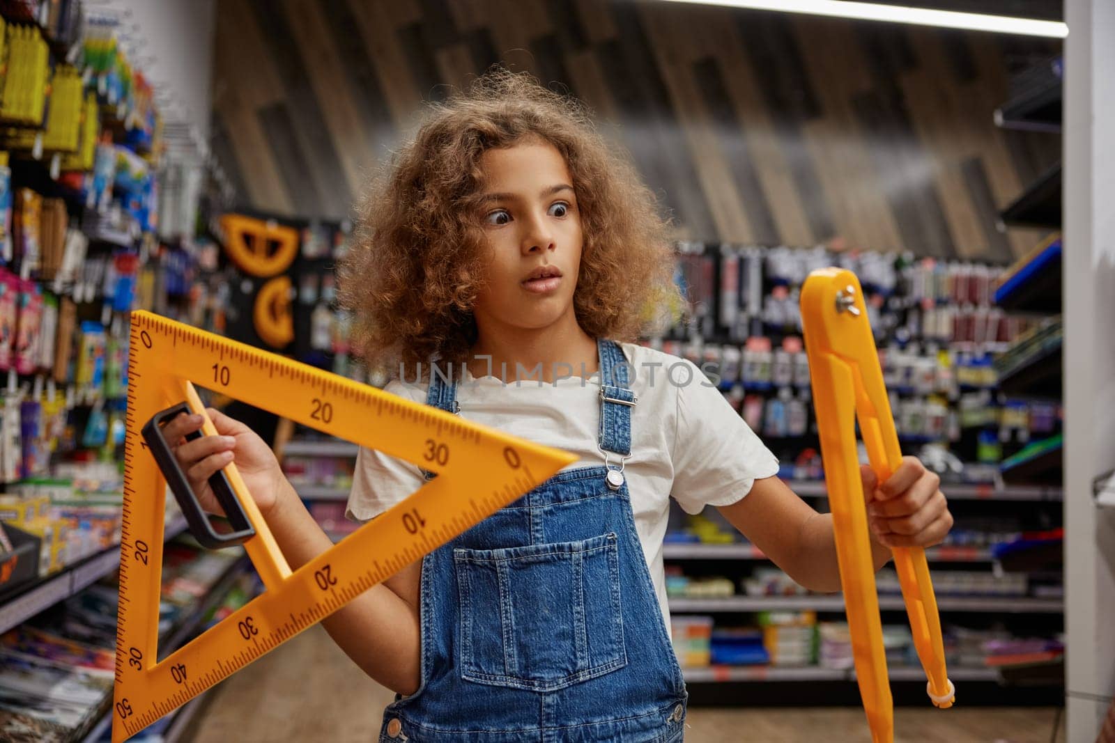 Shocked girl student holding huge compass and triangle ruler stationery in hands while standing at store. Creative studying tools for education