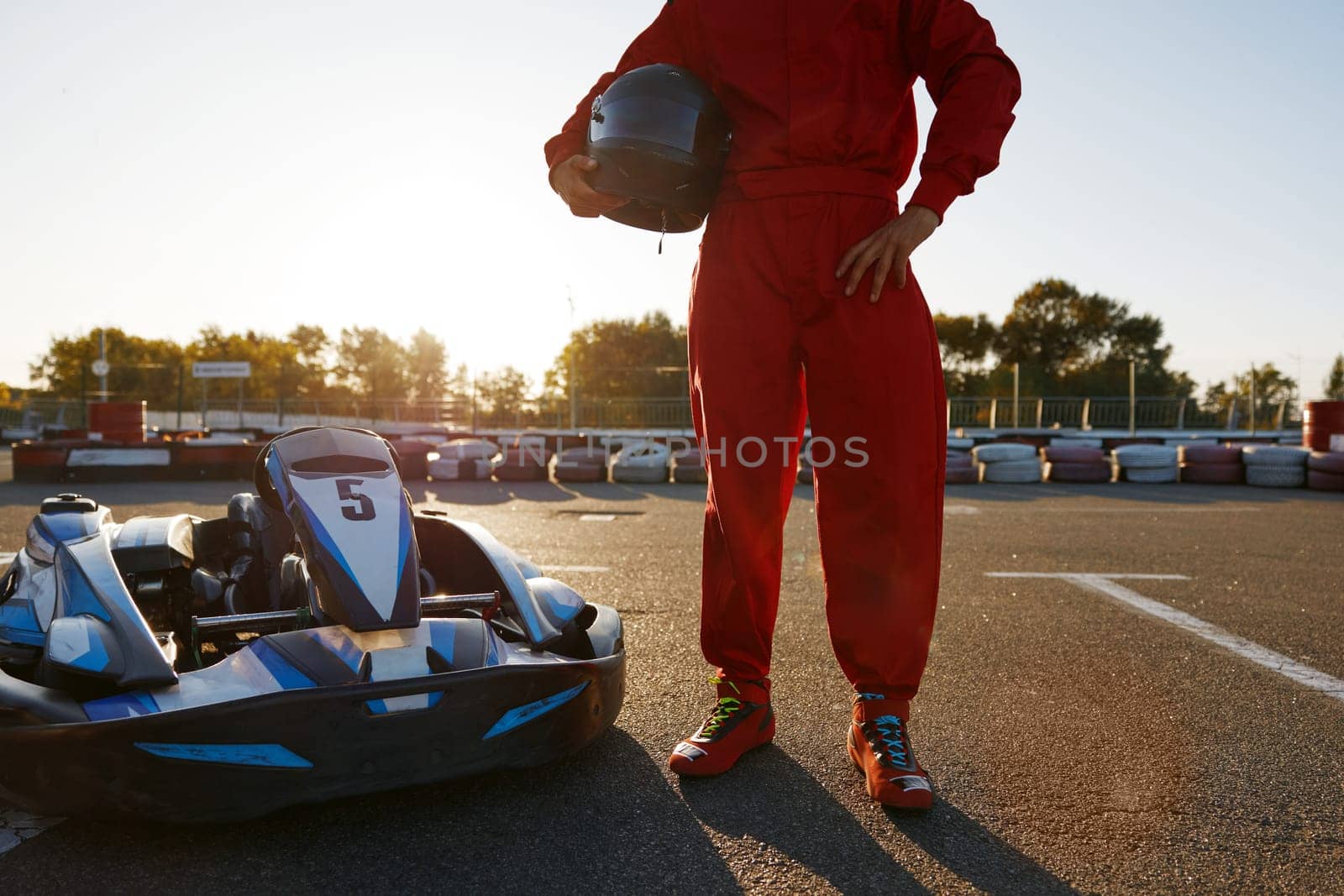 Cropped closeup shot of go-kart driver wearing overalls holding helmet in hands standing over motorsport training track
