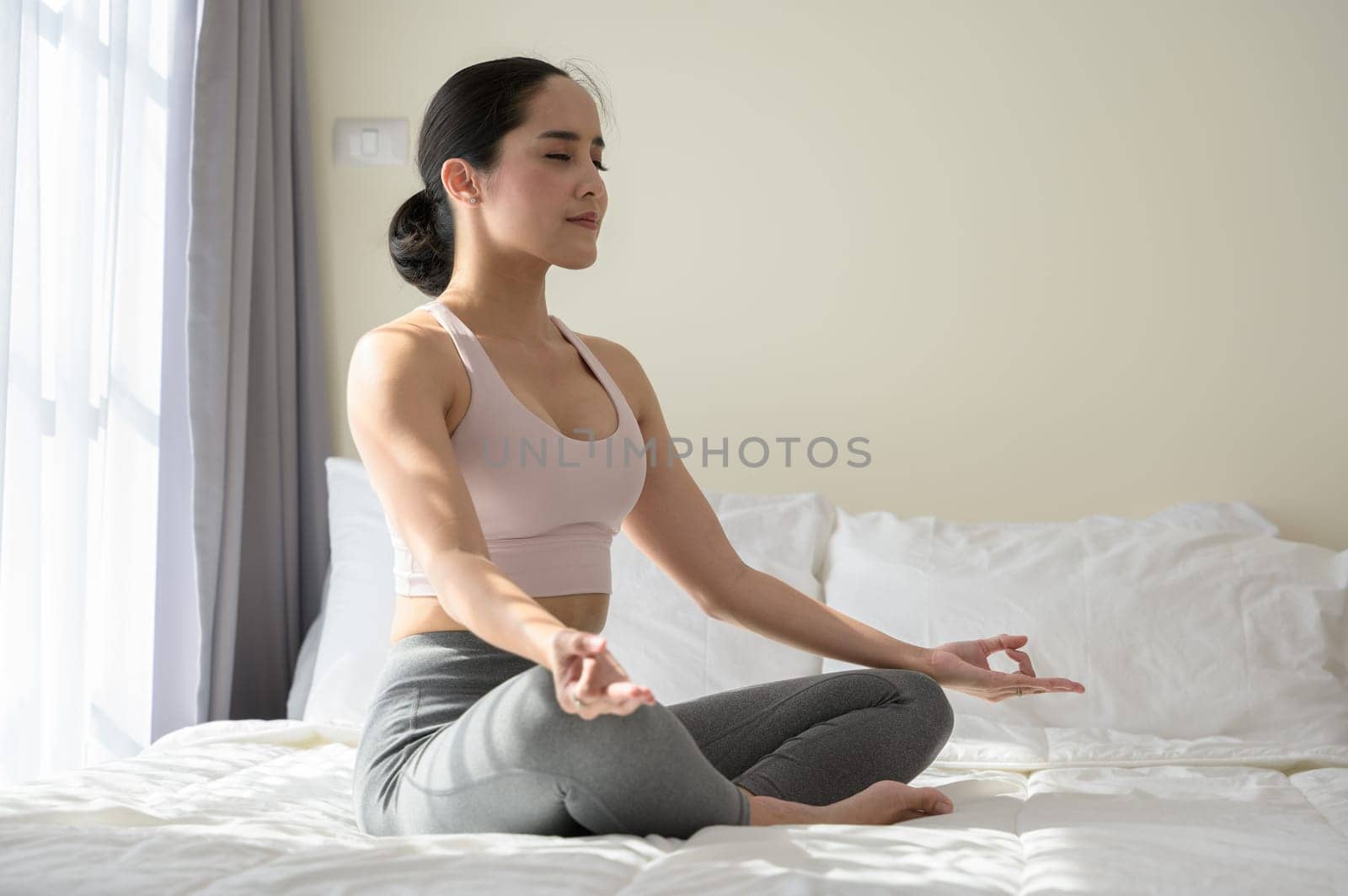 Asian female in sports clothes sitting in lotus position on bed. Woman doing yoga exercise meditating relaxed with closed eyes at home. Morning workout in bedroom. Healthy, relaxation and sport lifestyle.