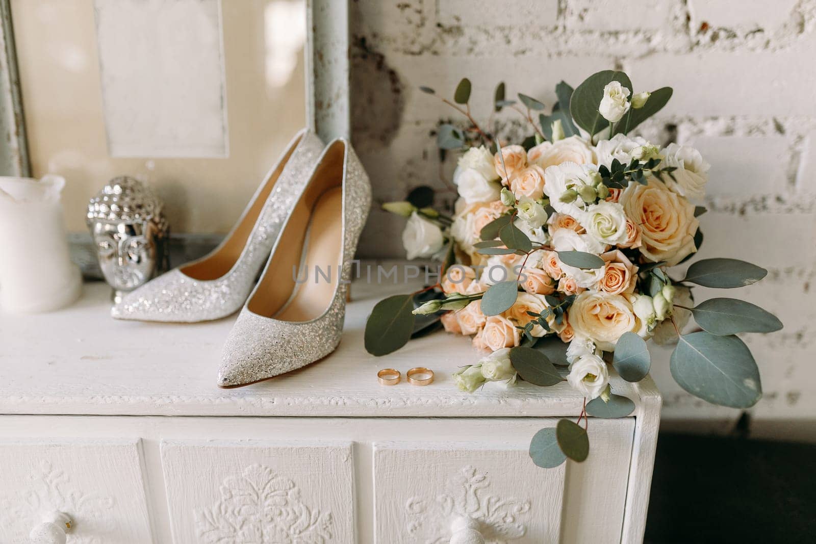 Wedding shoes and wedding paraphernalia, gold wedding rings, a wedding bouquet on a white chest of drawers near empty photo frames