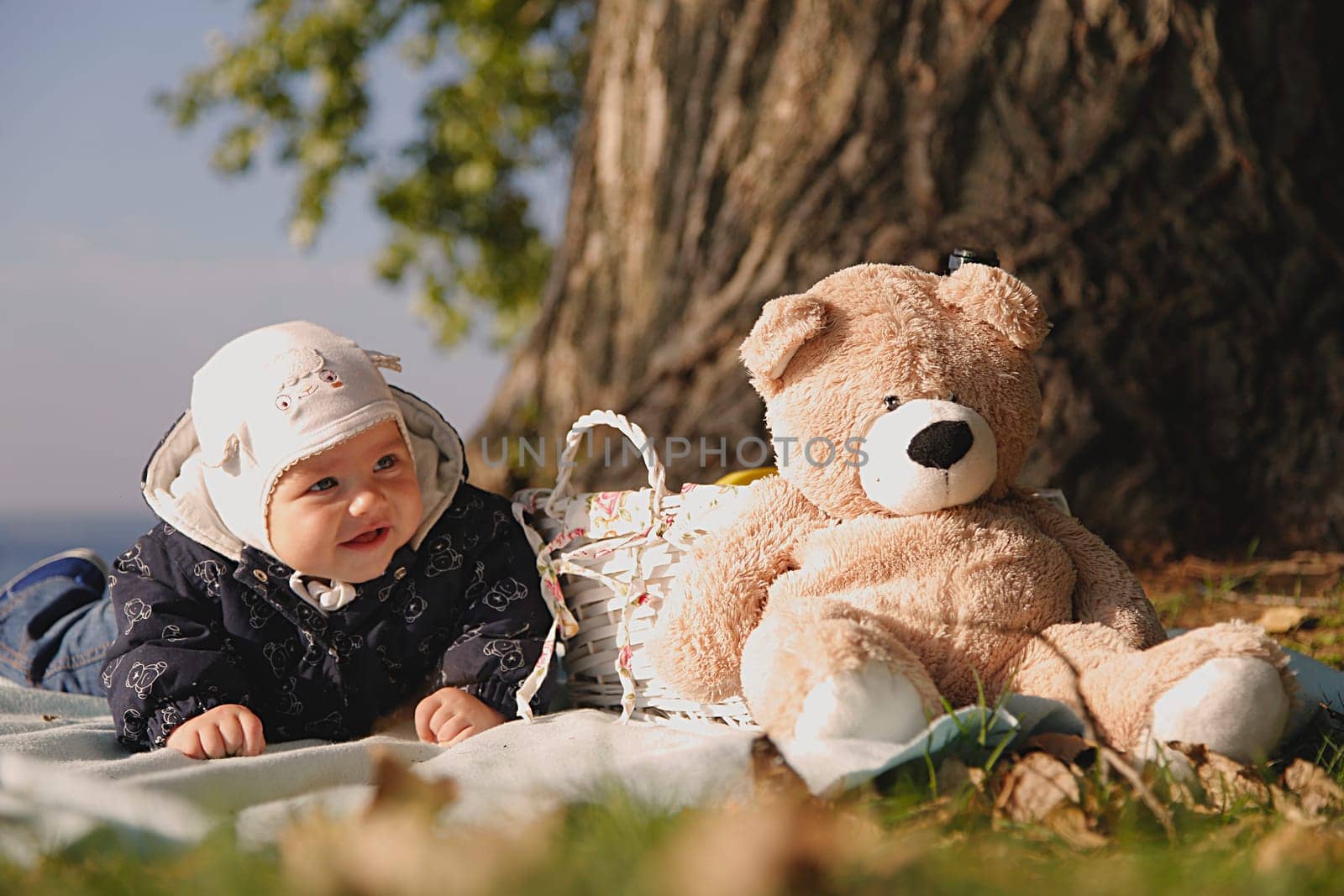 A little kid on a picnic with a teddy bear and a wicker basket near a large tree and the blue sea, a close-up view from below