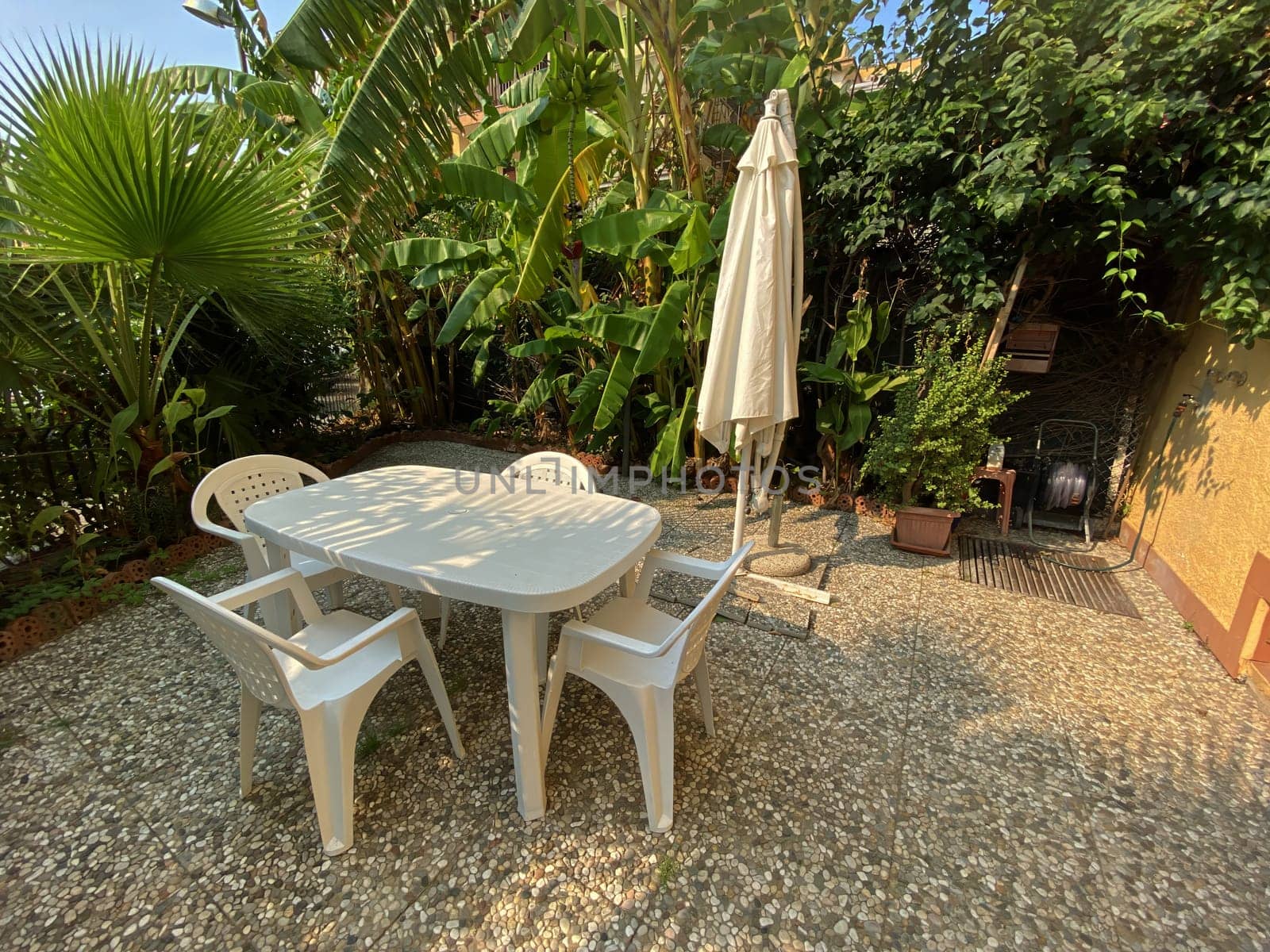 Cozy garden corner with white plastic furniture under an umbrella, surrounded by lush greenery for outdoor relaxation