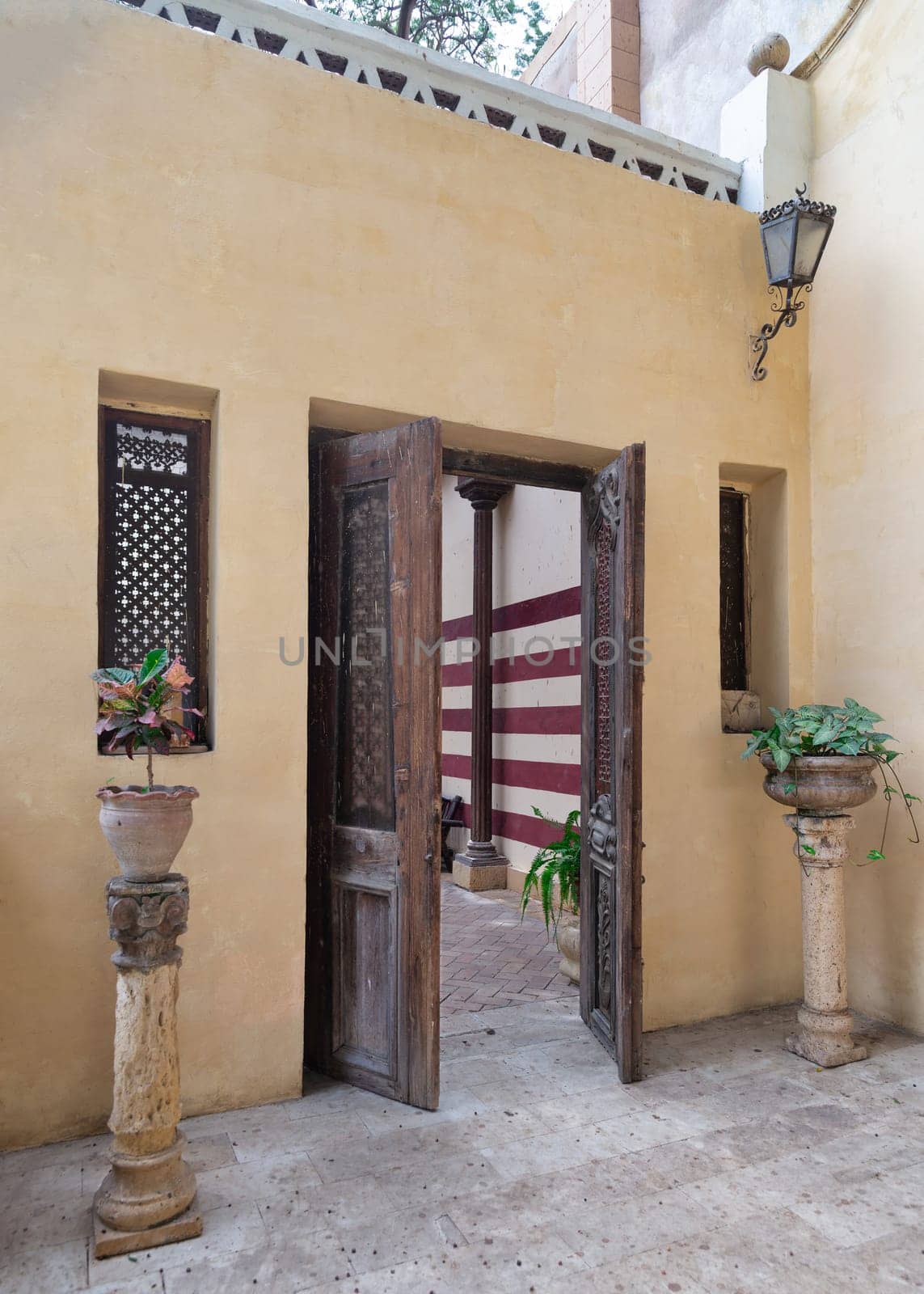 Open antique wooden decorated doorway mediating two wooden windows and leading to a peaceful courtyard with lush greenery and stone staircase