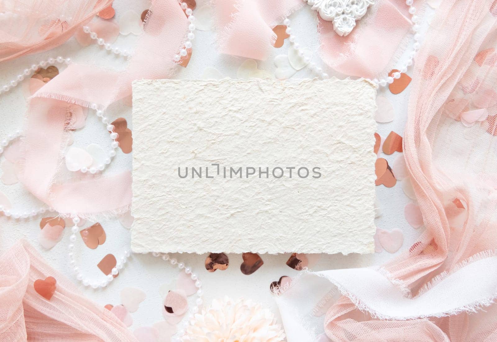 Handmade paper card near pink decorations, hearts and silk ribbons on white table top view, mockup. Romantic scene with vertical blank card flat lay. Wedding, Valentines, Spring stationery