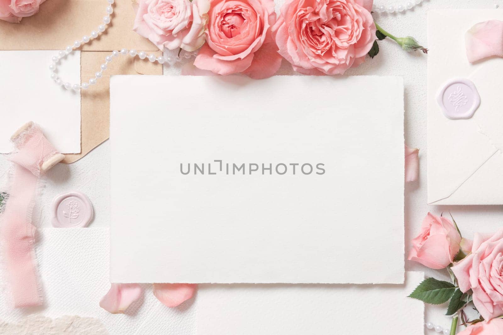 Blank card near light pink roses, buds, petals and silk ribbons on white table top view, wedding mockup. Romantic flat lay with horizontal card and pastel decor