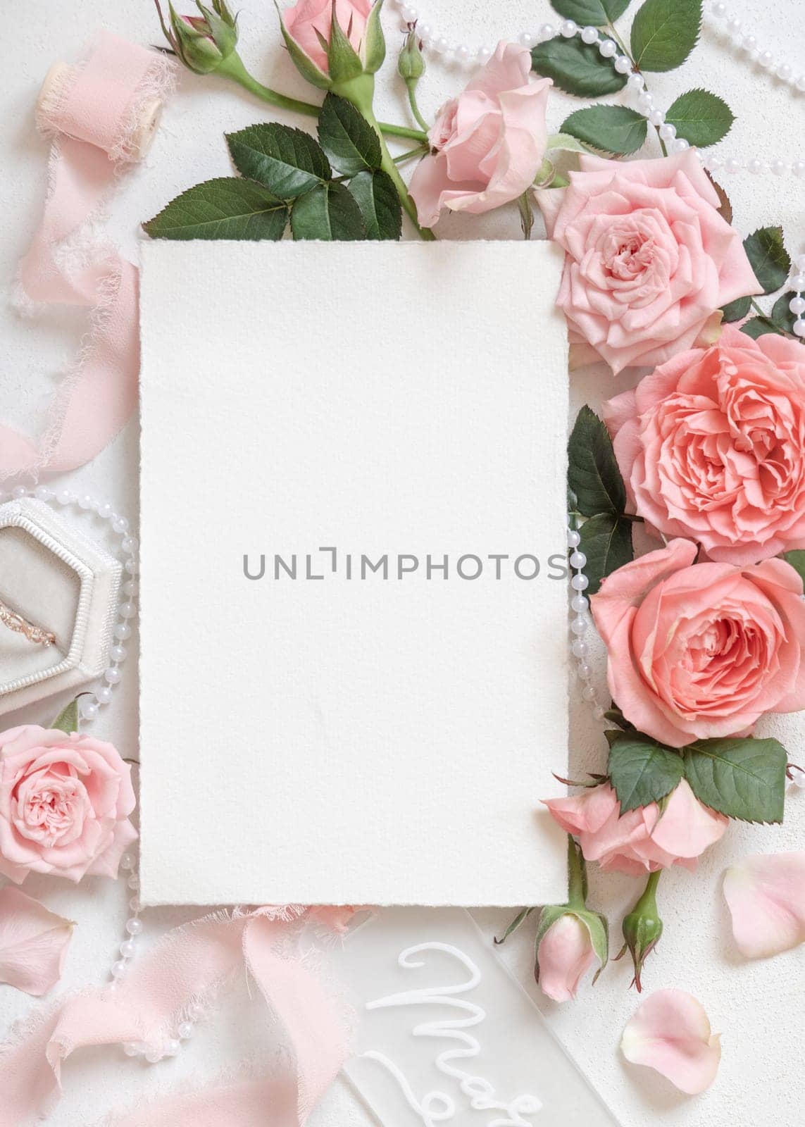Blank card near light pink roses, engagement ring in a box and silk ribbons on white table top view, wedding mockup. Romantic flat lay with Vertical card and pastel decor