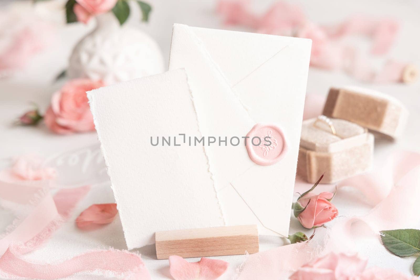 Card and sealed envelope near light pink roses, petals, engagement ring in a box and silk ribbons on white table close up, wedding mockup. Romantic scene with pastel decor