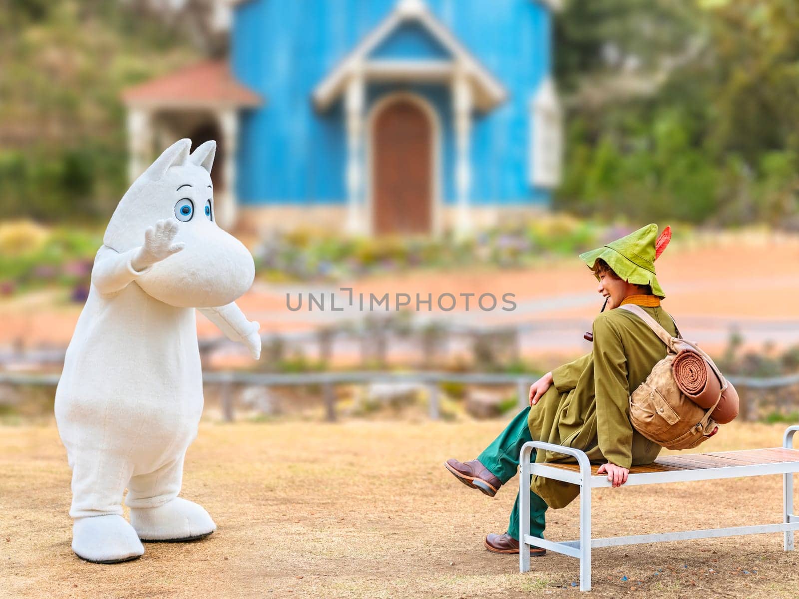 japan, saitama - mar 26 2023: Moomintroll costume waving hand to his smiling friend Snufkin wearing a green hat carrying a backpack with a camp mattress smoking a pipe on a bench in Moominvalley park.